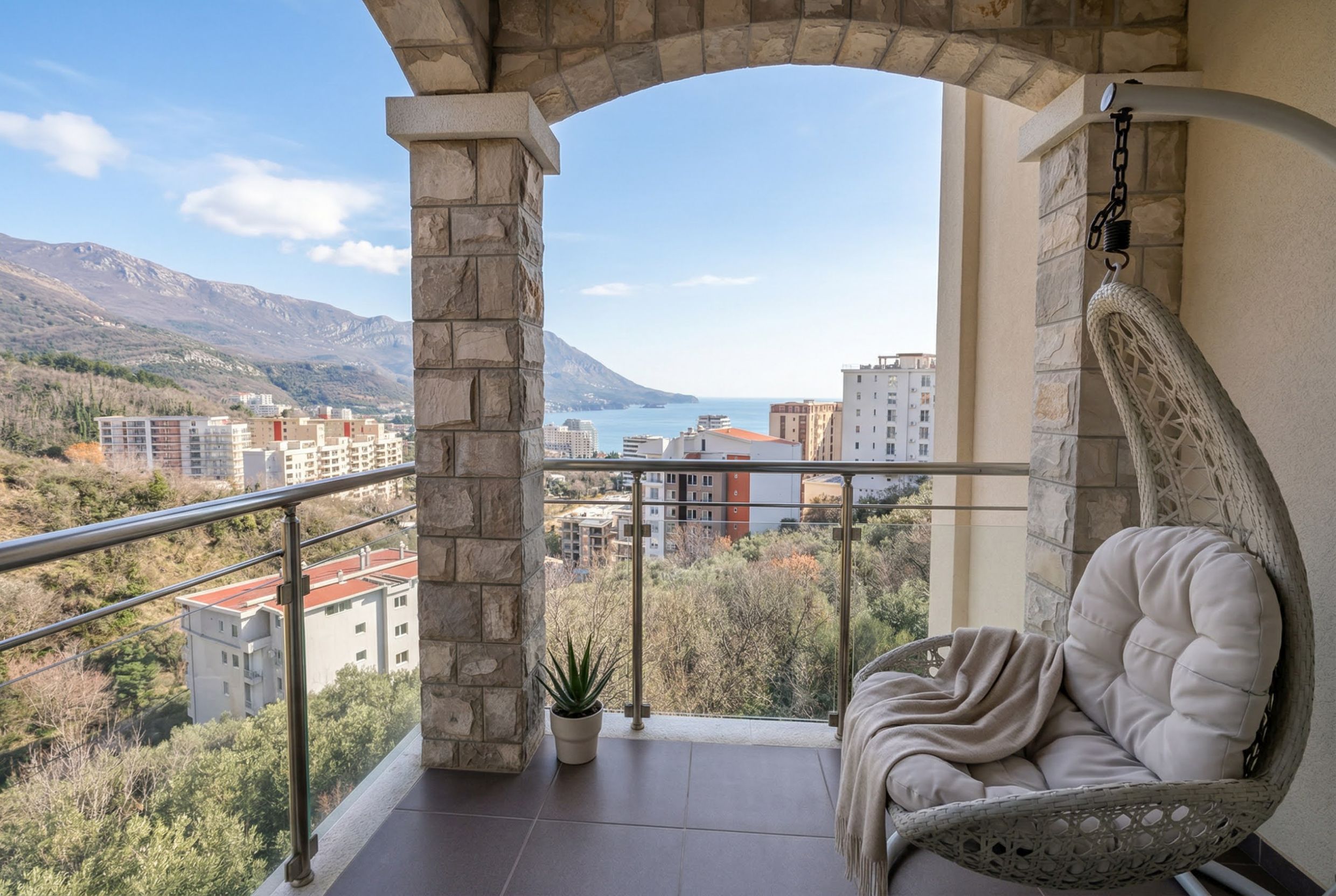 Balcony with a hanging egg chair and a view of the sea and mountains in an apartment in Bečići, Montenegro. Stone-clad columns.