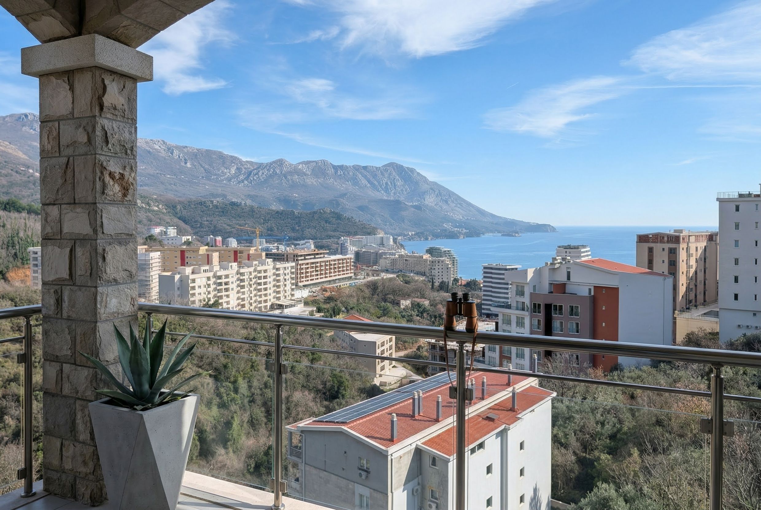View from the apartment balcony in Bečići, Montenegro: a panorama of mountains, sea, and city, with stone finishes and glass railings.