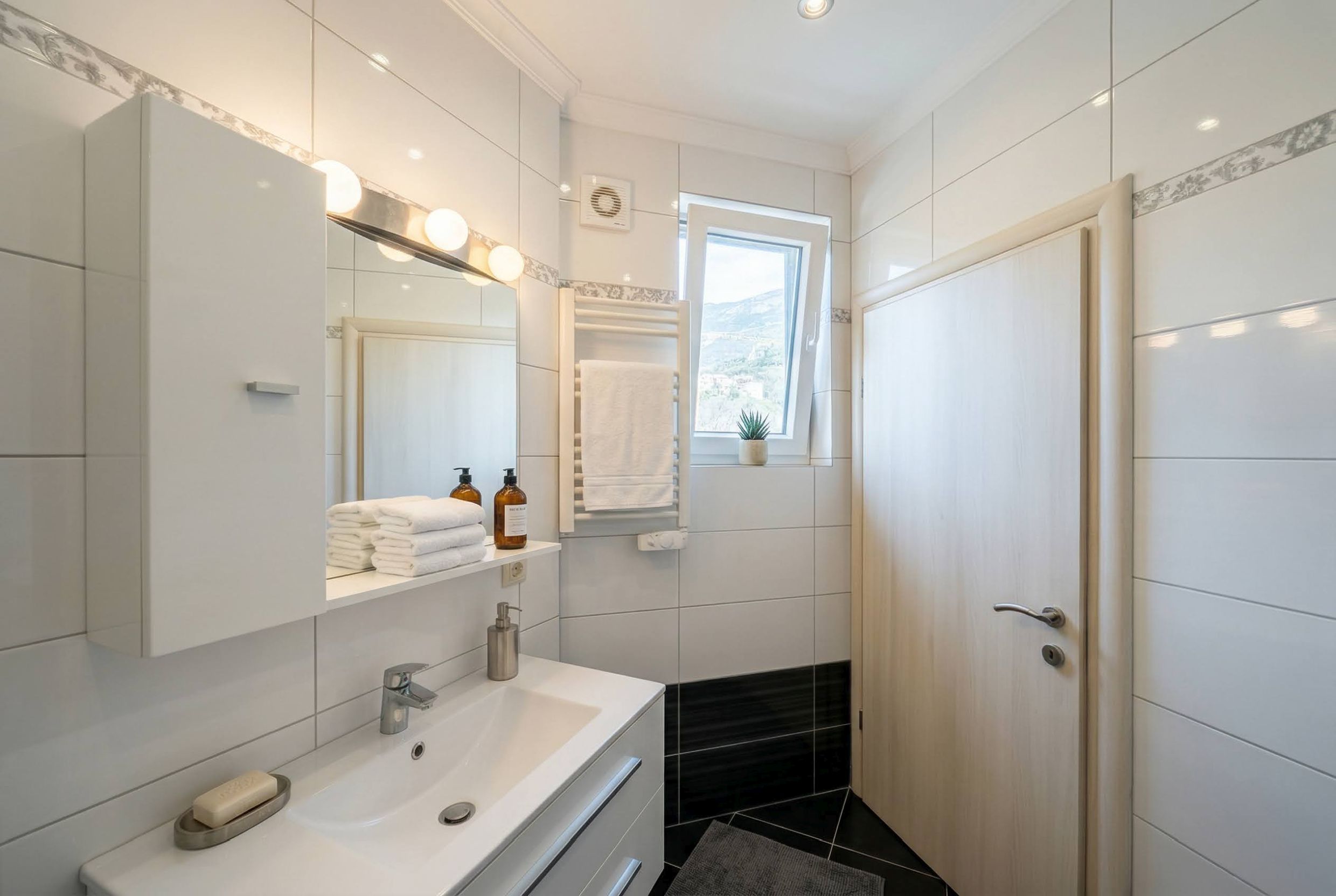 Bright bathroom in an apartment in Bečići, Montenegro: white tiles, modern fixtures, and a mountain view from the window.