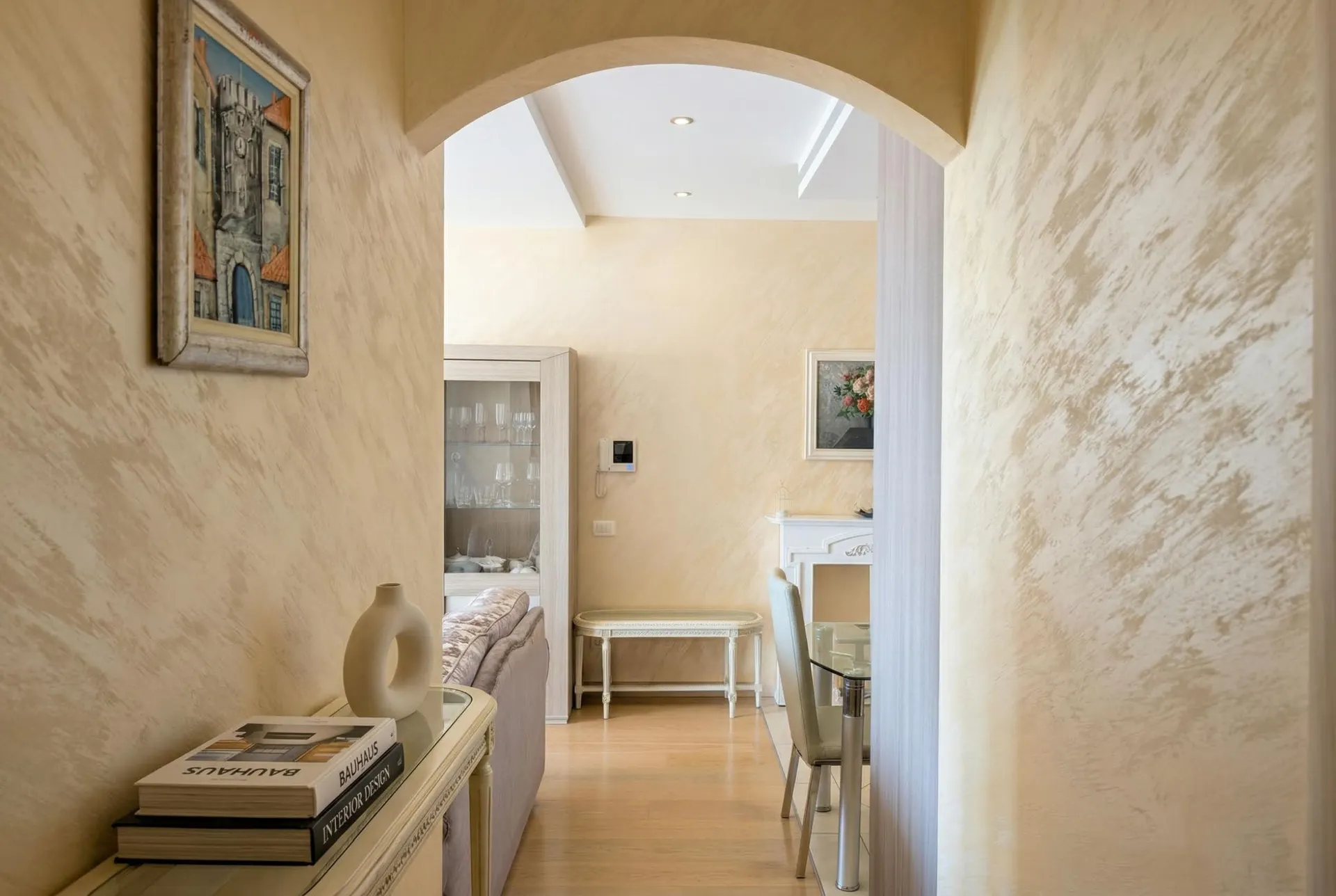 A hallway with an arch leads to a bright living room featuring beige textured walls, a sofa, and a glass display cabinet.