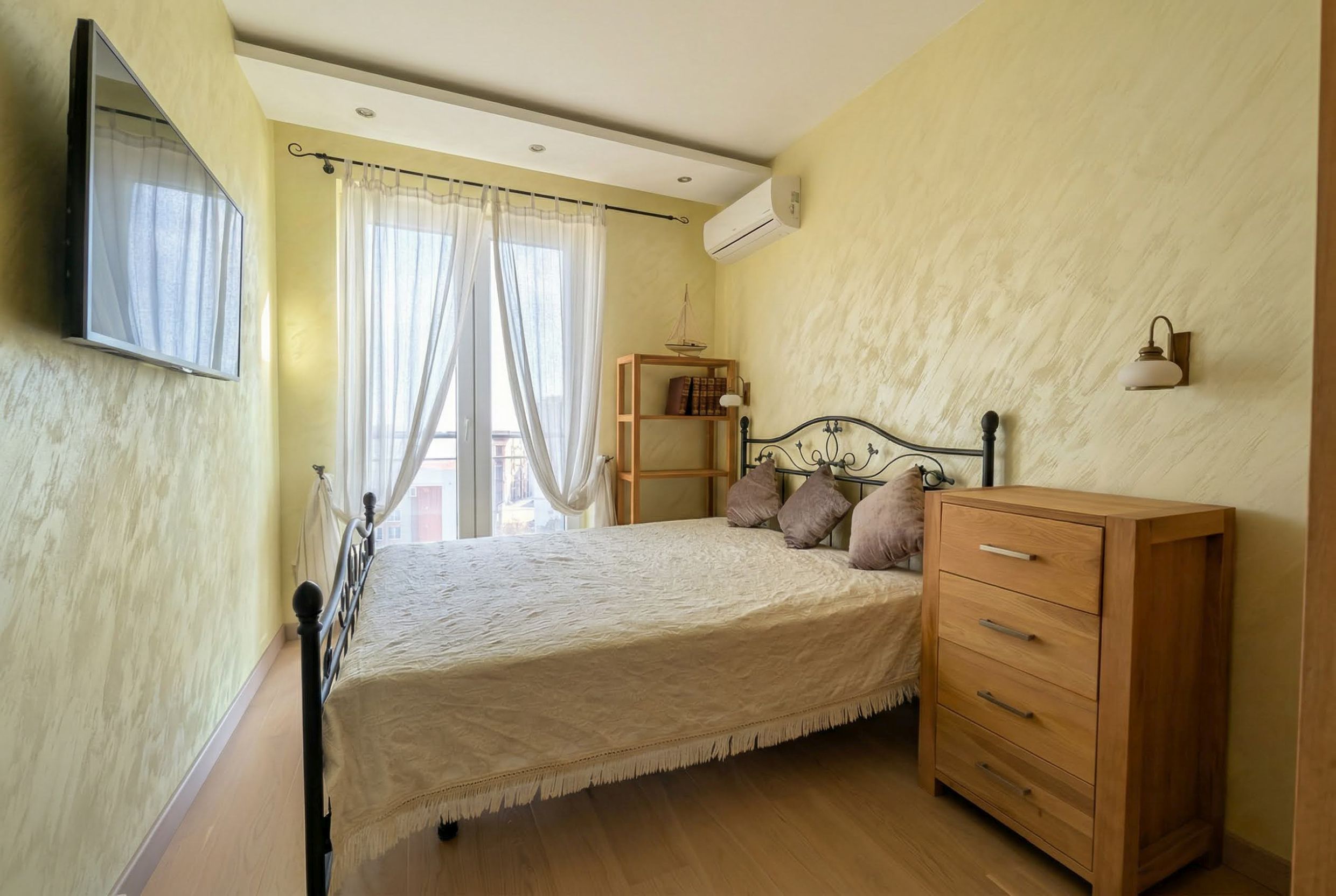 Cozy bedroom with a wrought-iron bed and wooden dresser in an apartment, Bečići, Montenegro. Light walls and access to the balcony.