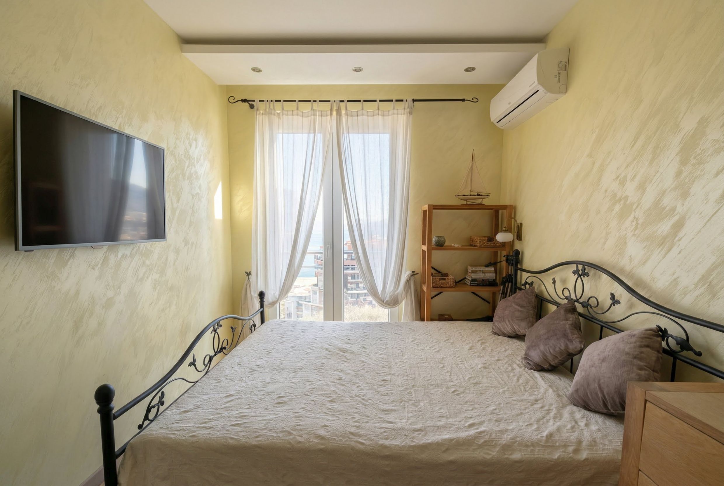 Bedroom with a wrought-iron bed and sea view in an apartment in Bečići, Montenegro. Light walls, TV, and access to the balcony.