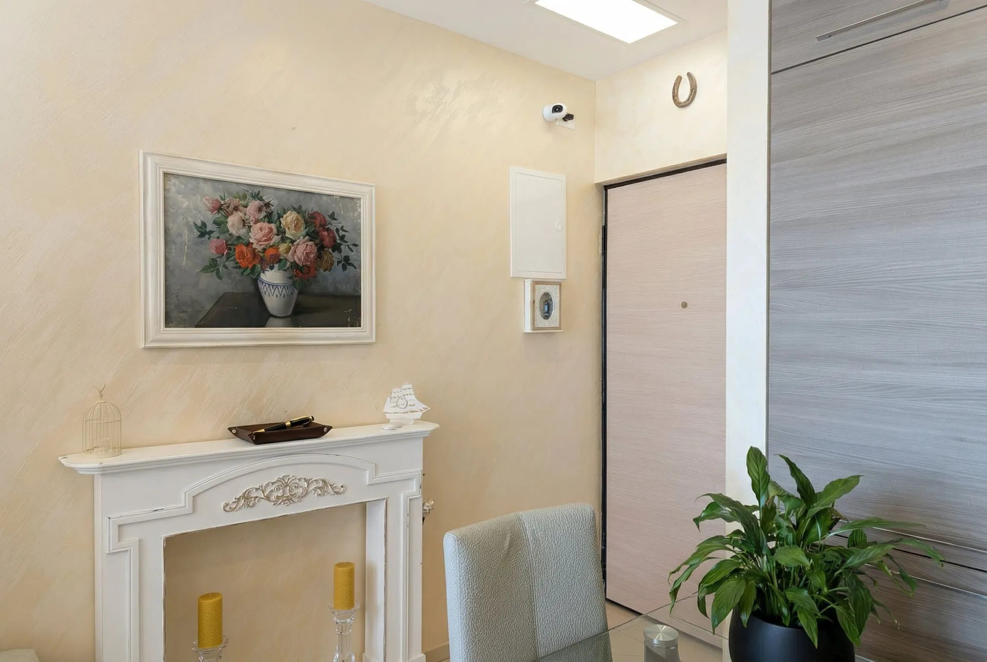 Living room interior featuring a white decorative fireplace, floral painting, and entrance door.