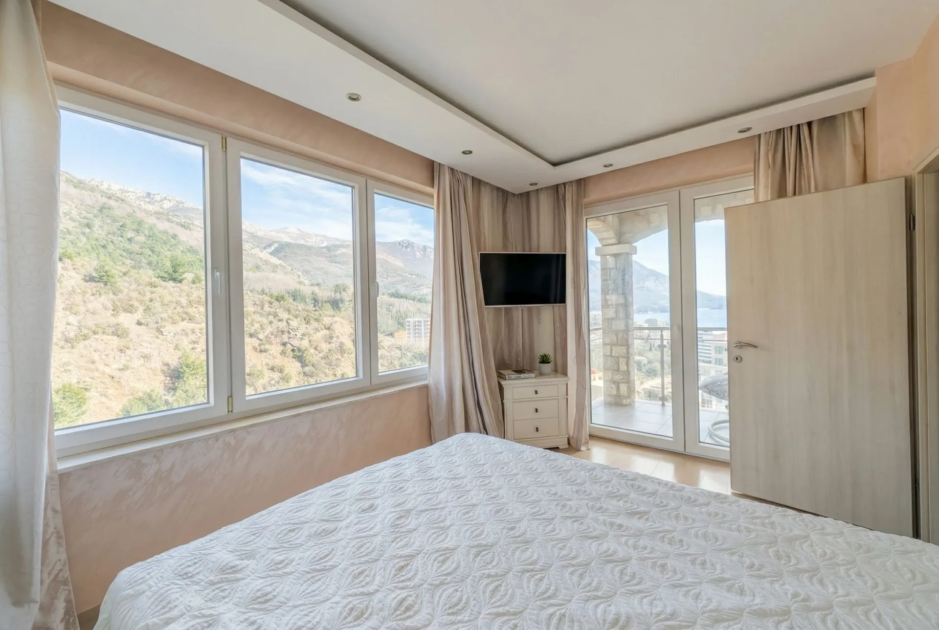 Bright bedroom with white bed, wall-mounted TV, and large windows offering a scenic view of mountains and sea.