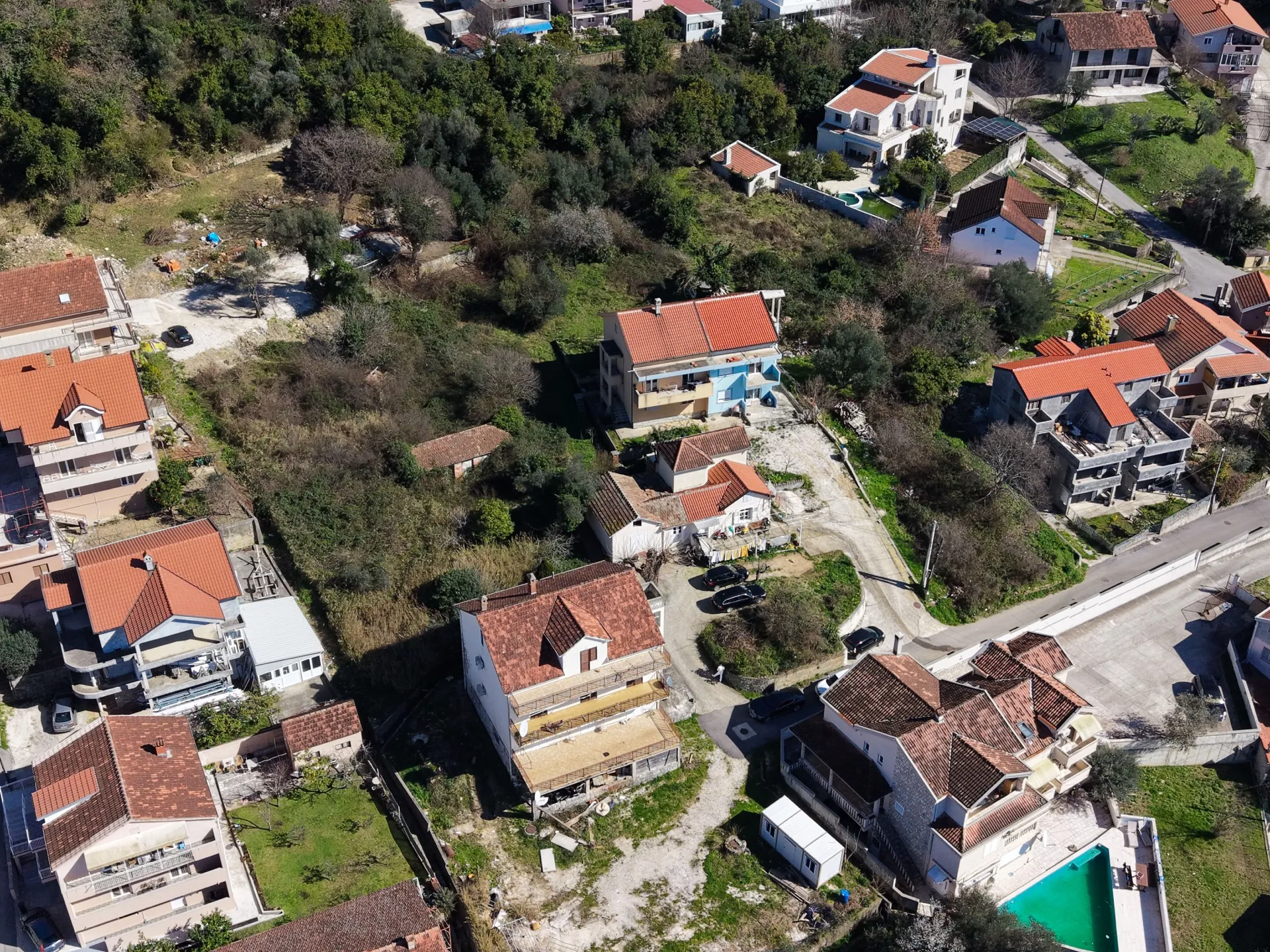 Aerial view of a residential area featuring houses with red tile roofs, green hills, and private swimming pools.