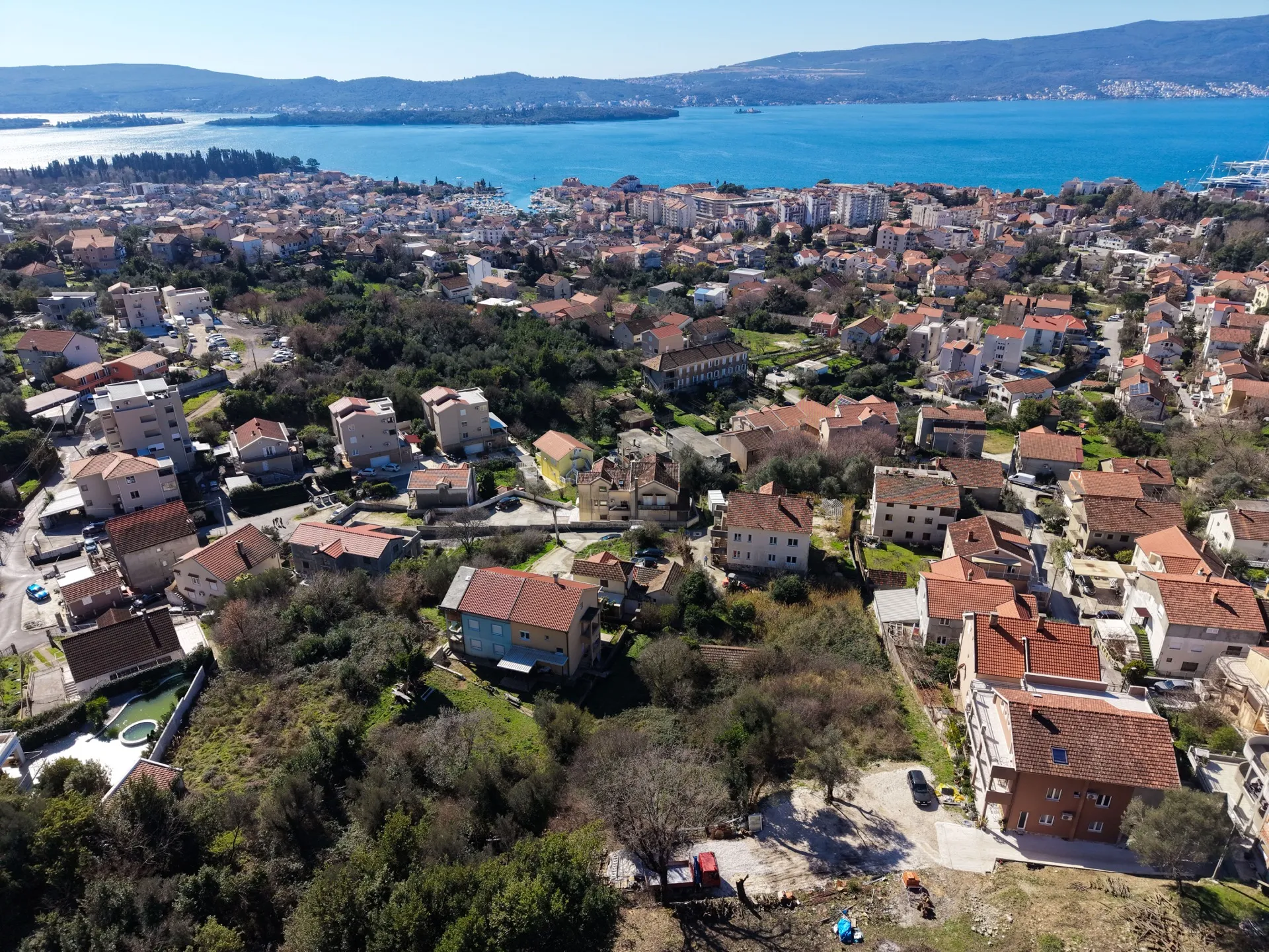 Aerial view of a coastal town with houses featuring red tiled roofs surrounded by greenery, set against a backdrop of blue sea and mountains.