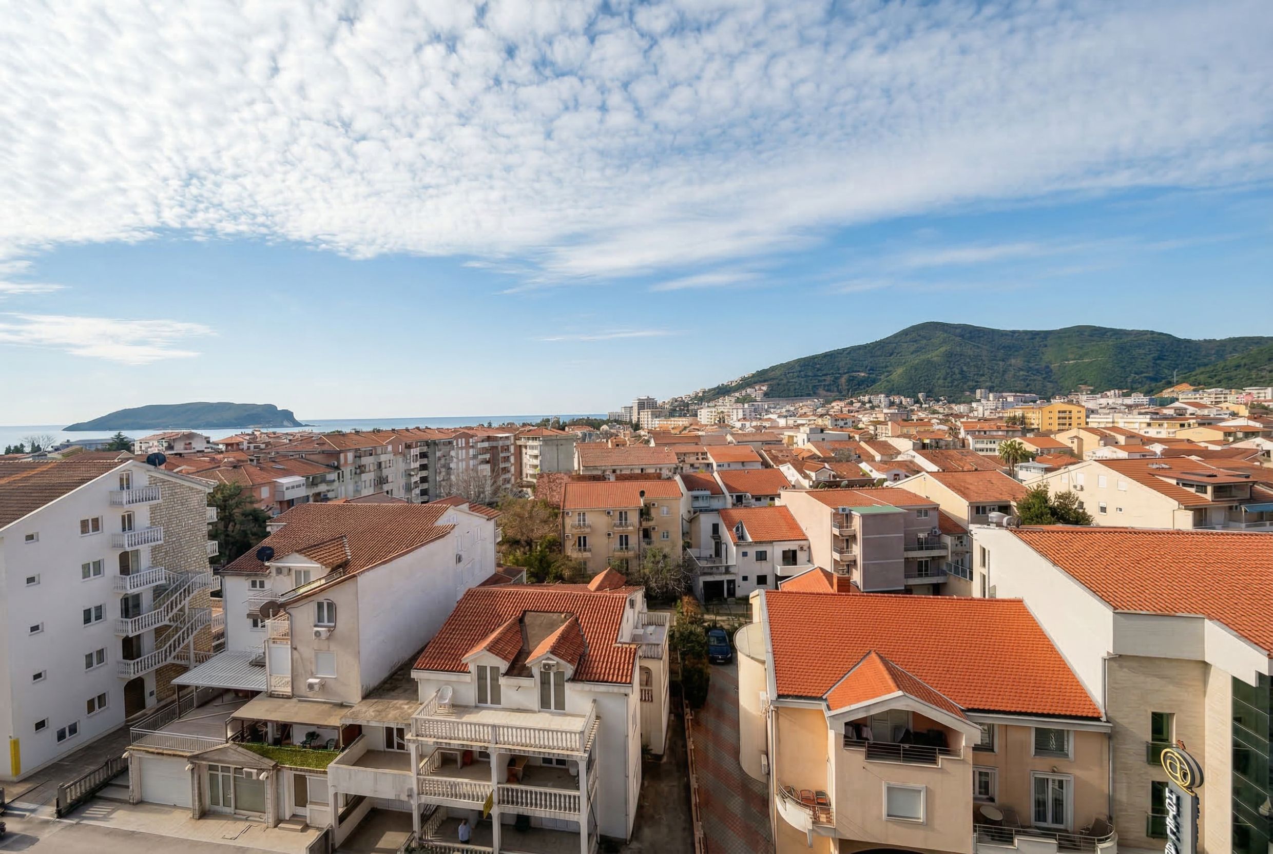 Panoramic view of Budva city and the Adriatic Sea from a penthouse in Montenegro, featuring terracotta roofs and mountains.