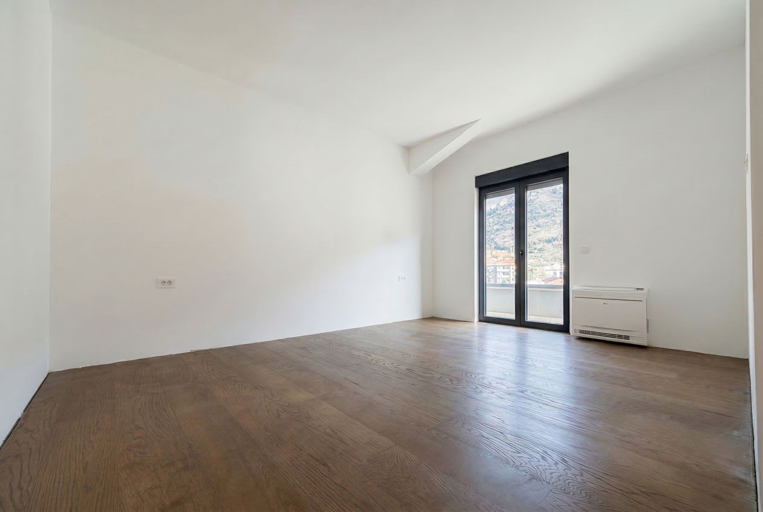 Bright living room in a penthouse in Budva, Montenegro: parquet flooring, white walls, panoramic door, and mountain view.