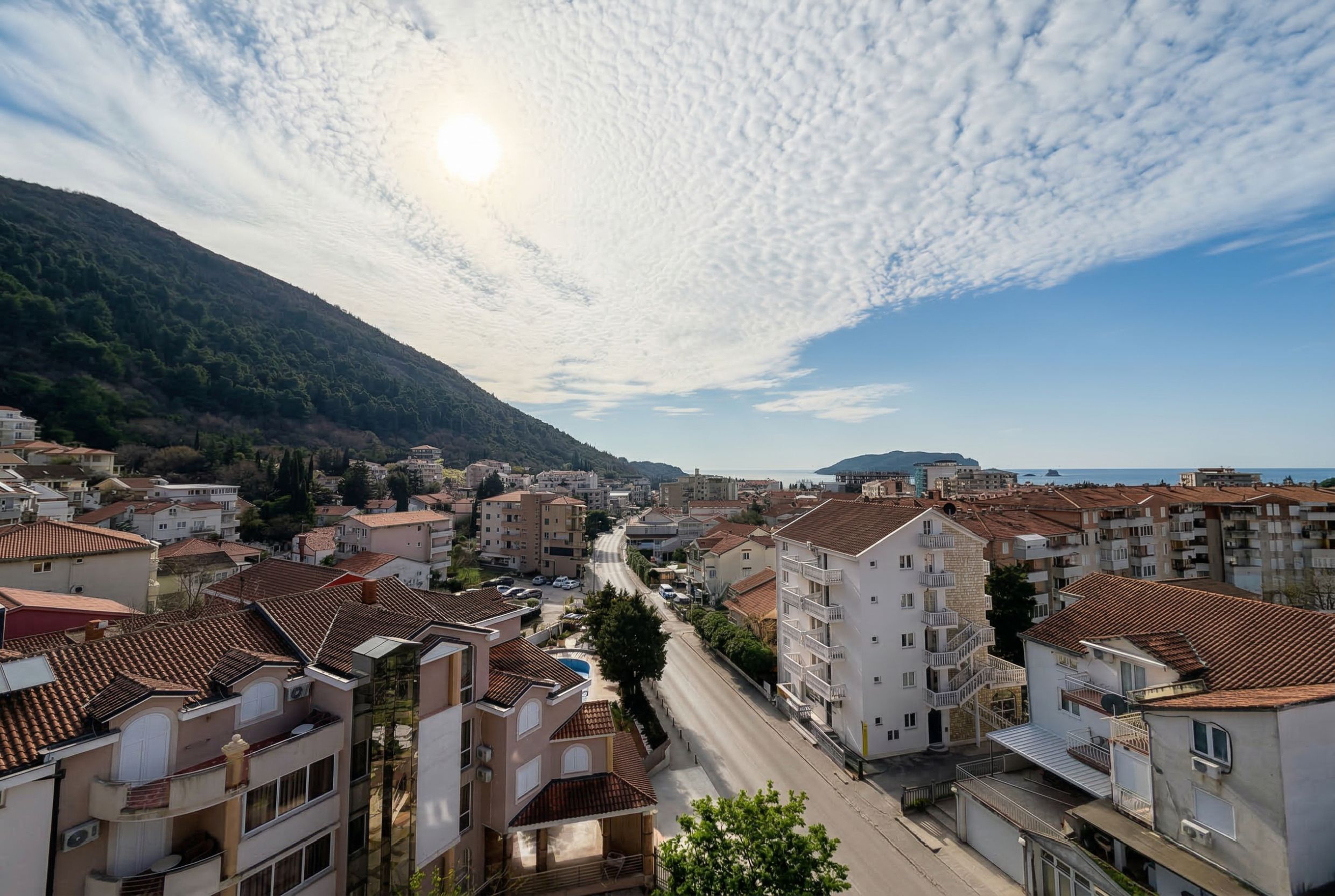 Panoramic view of Budva, Montenegro: residential buildings with terracotta roofs, mountains, and the sea horizon from a terrace.