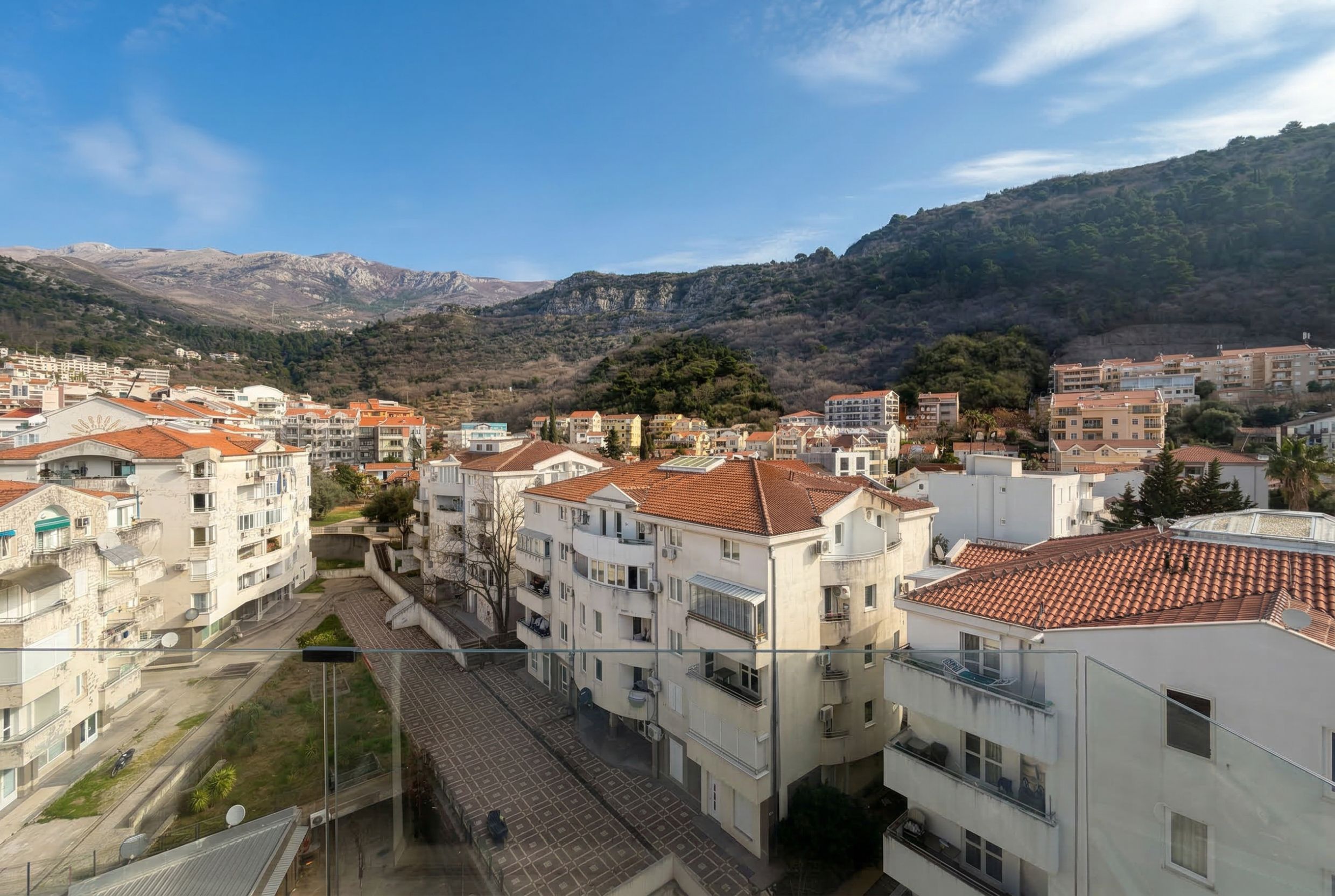 View from the penthouse balcony in Budva, Montenegro: panoramic view of the city, residential buildings with tiled roofs, and mountain landscape.