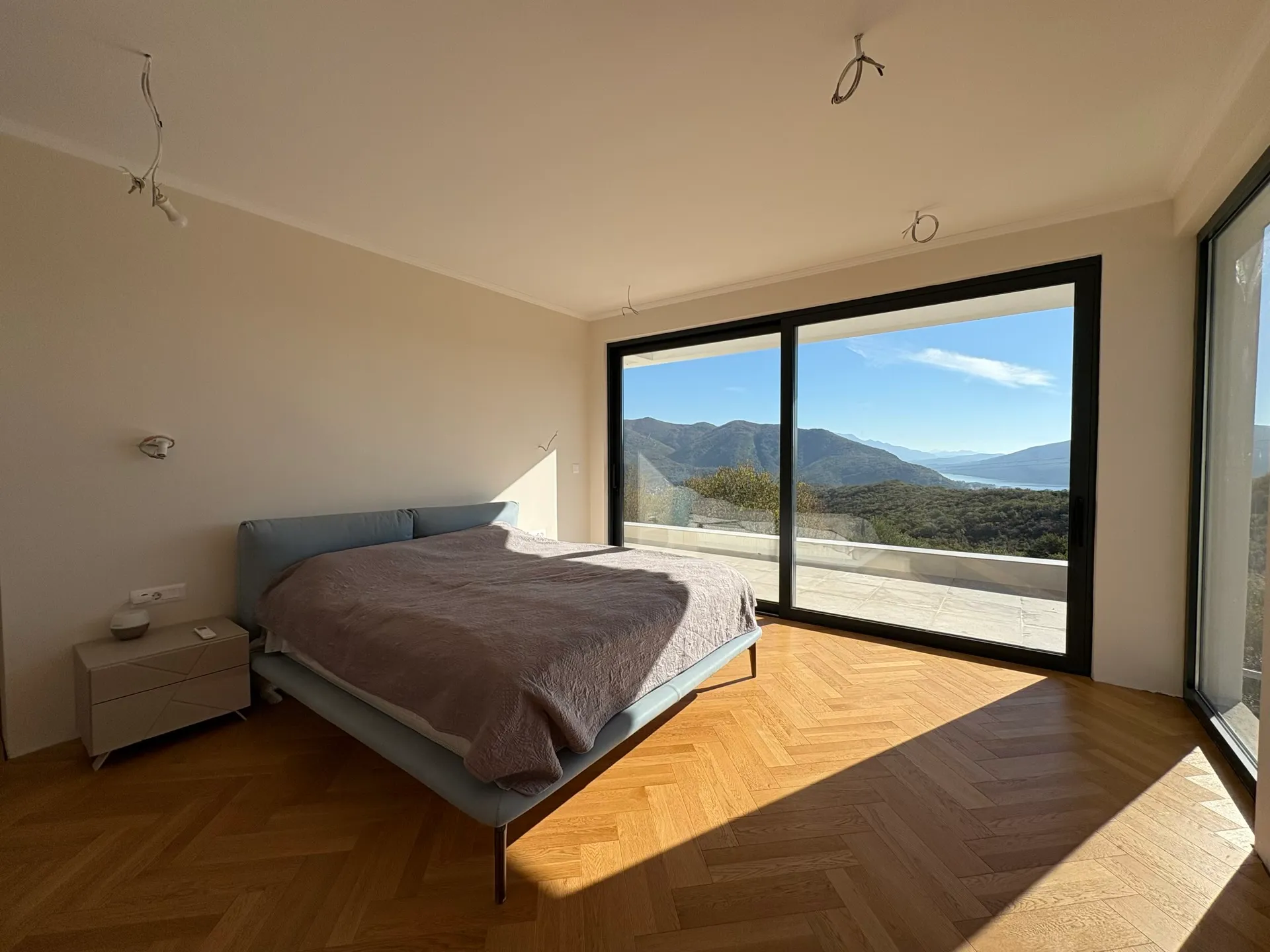 Bright bedroom with a double bed, herringbone wood floor, and large windows overlooking mountains.