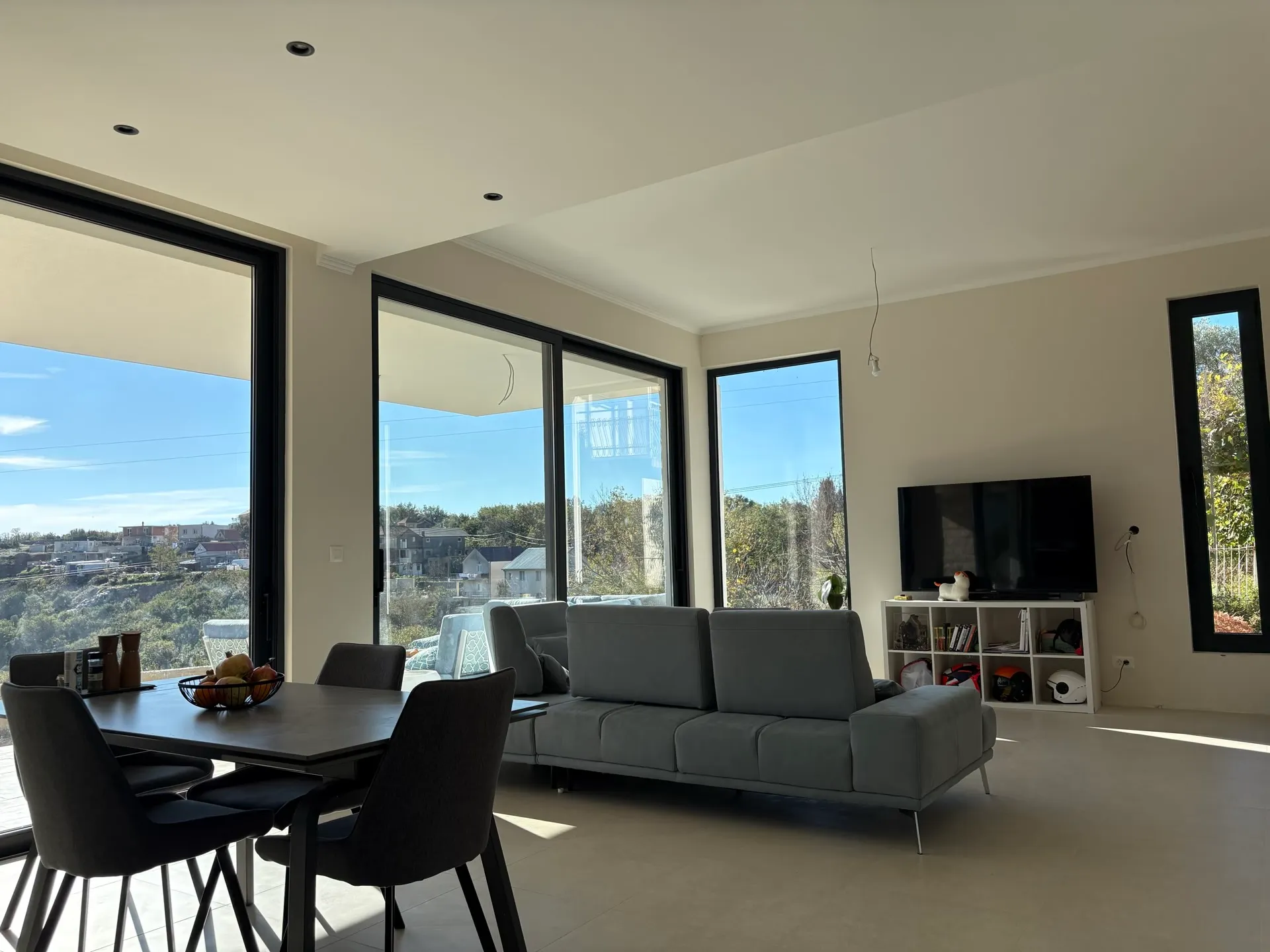 Bright room featuring a grey sofa, dining table with chairs, and a TV on a white shelf near large windows.