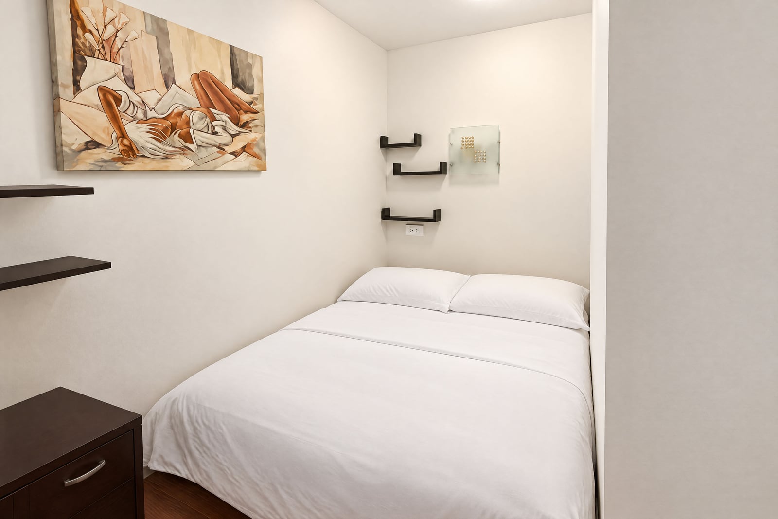 Small bedroom with a white bed, dark nightstand, and artwork on the wall.