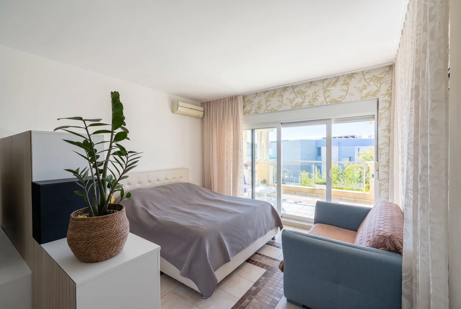 Bright bedroom featuring a double bed, blue sofa, potted plant, and sliding glass door to balcony.