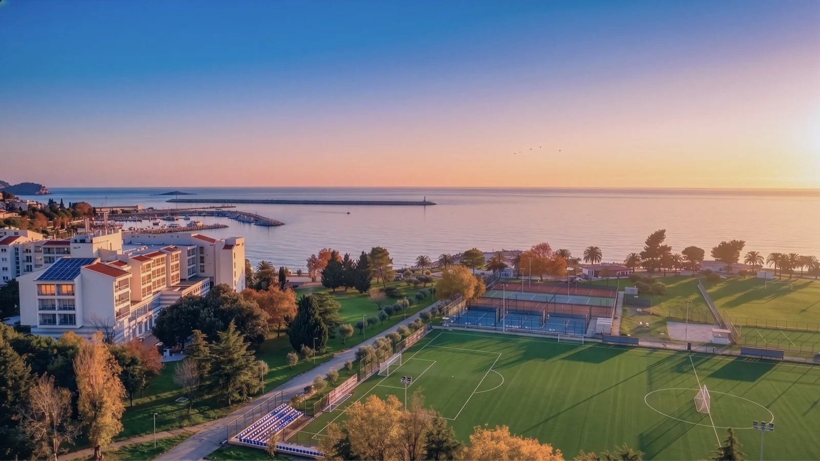 Aerial view of a sports complex with a soccer field and tennis courts near the sea at sunset.