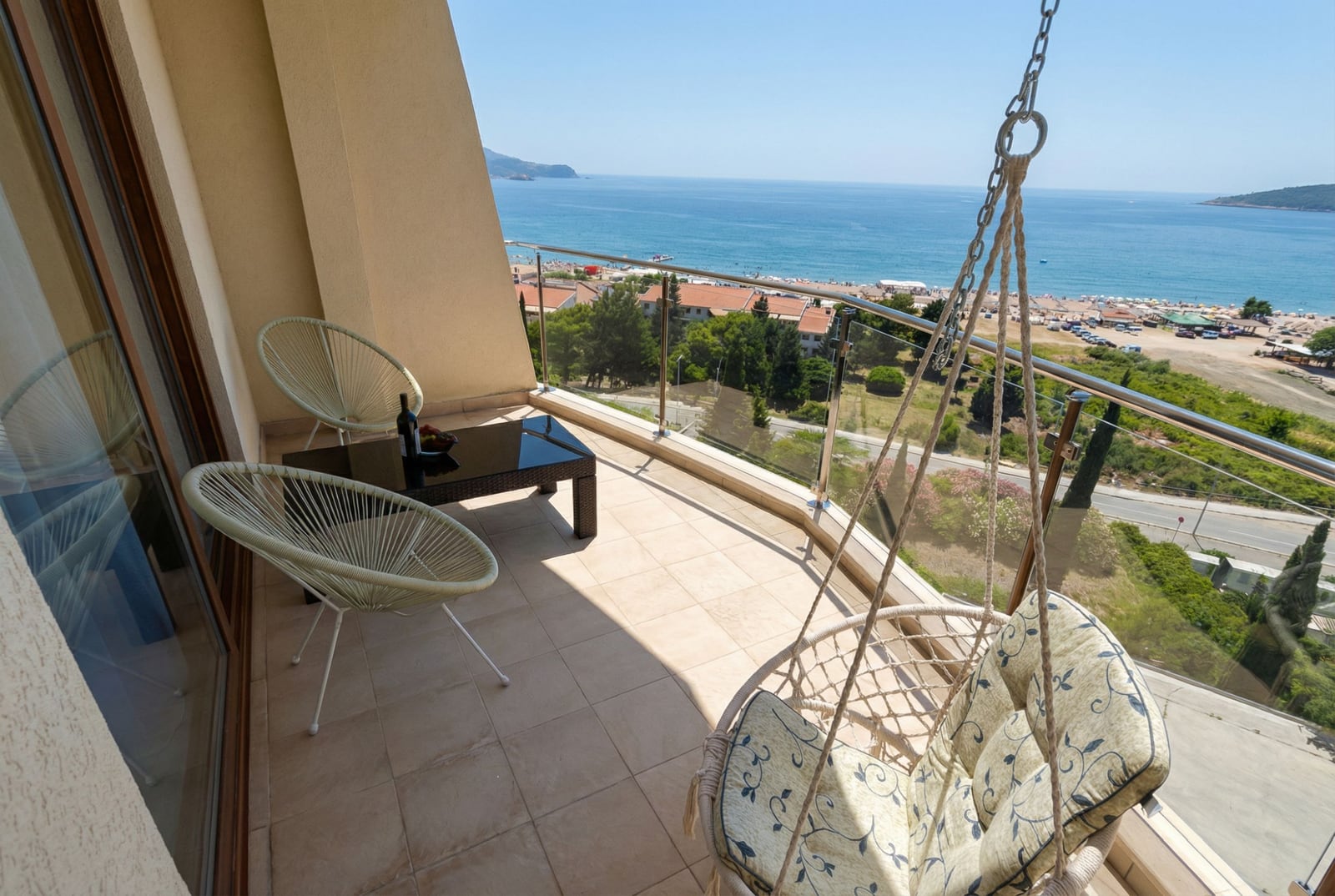 Apartment balcony in Bečići, Montenegro: wicker furniture, a hanging chair, and a panoramic view of the sea and beach.