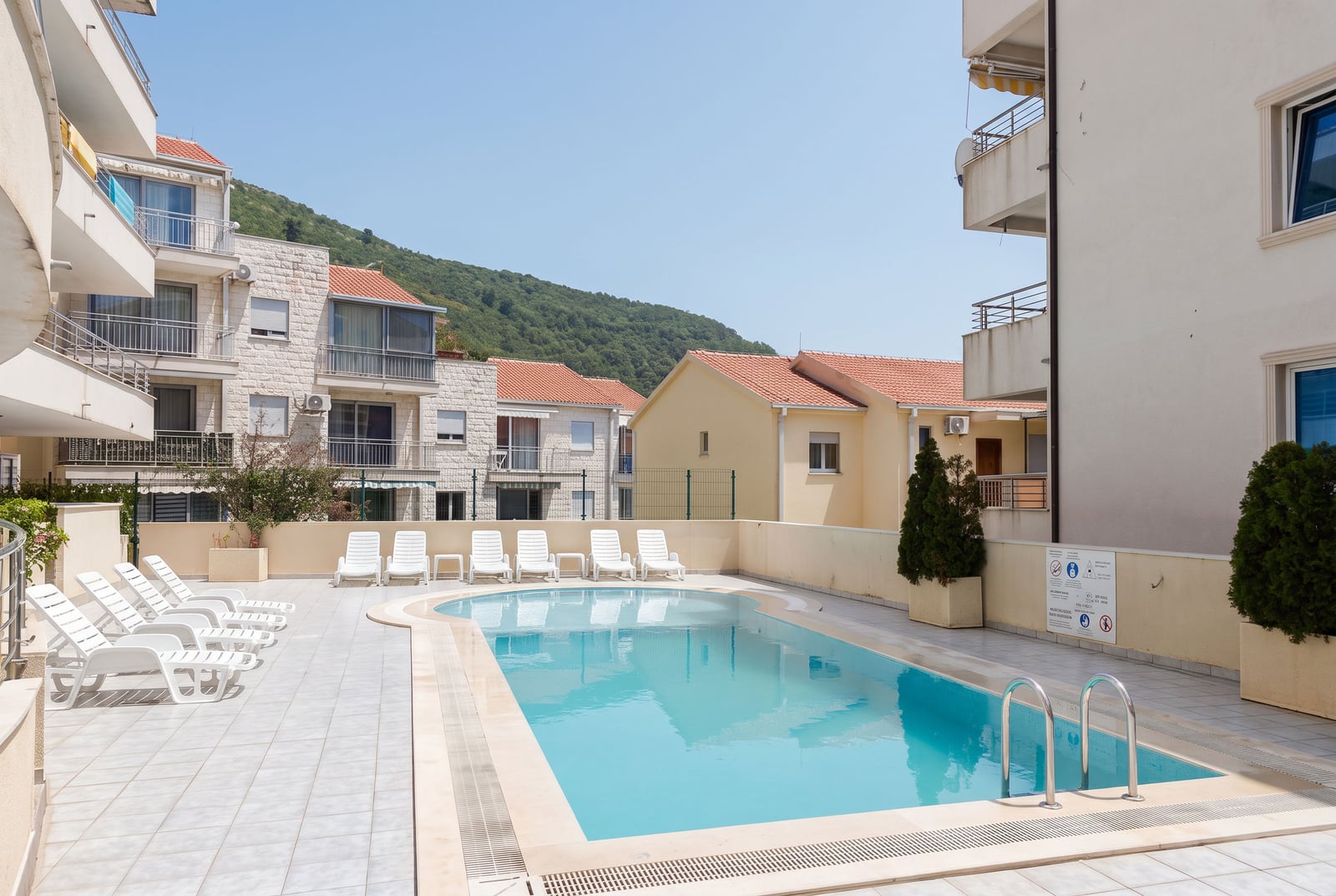 Outdoor swimming pool with white sun loungers on a tiled terrace surrounded by residential buildings.