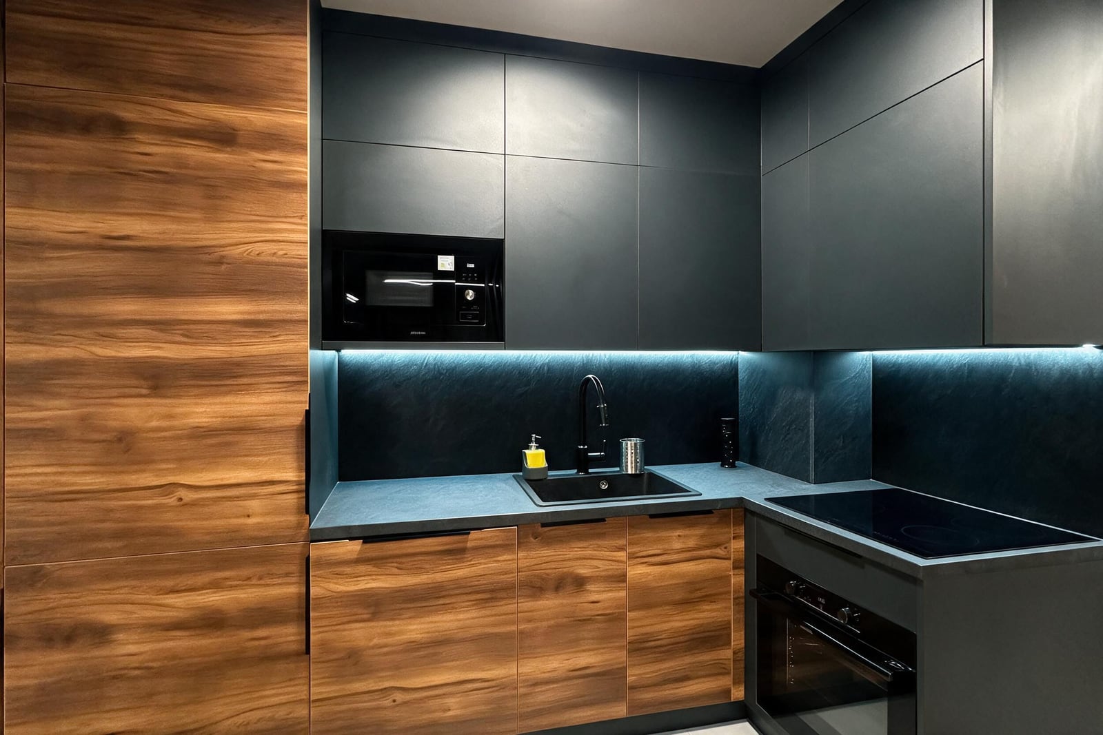 Modern kitchen with dark and wood cabinets, built-in microwave, cooktop, sink, and under-cabinet lighting.