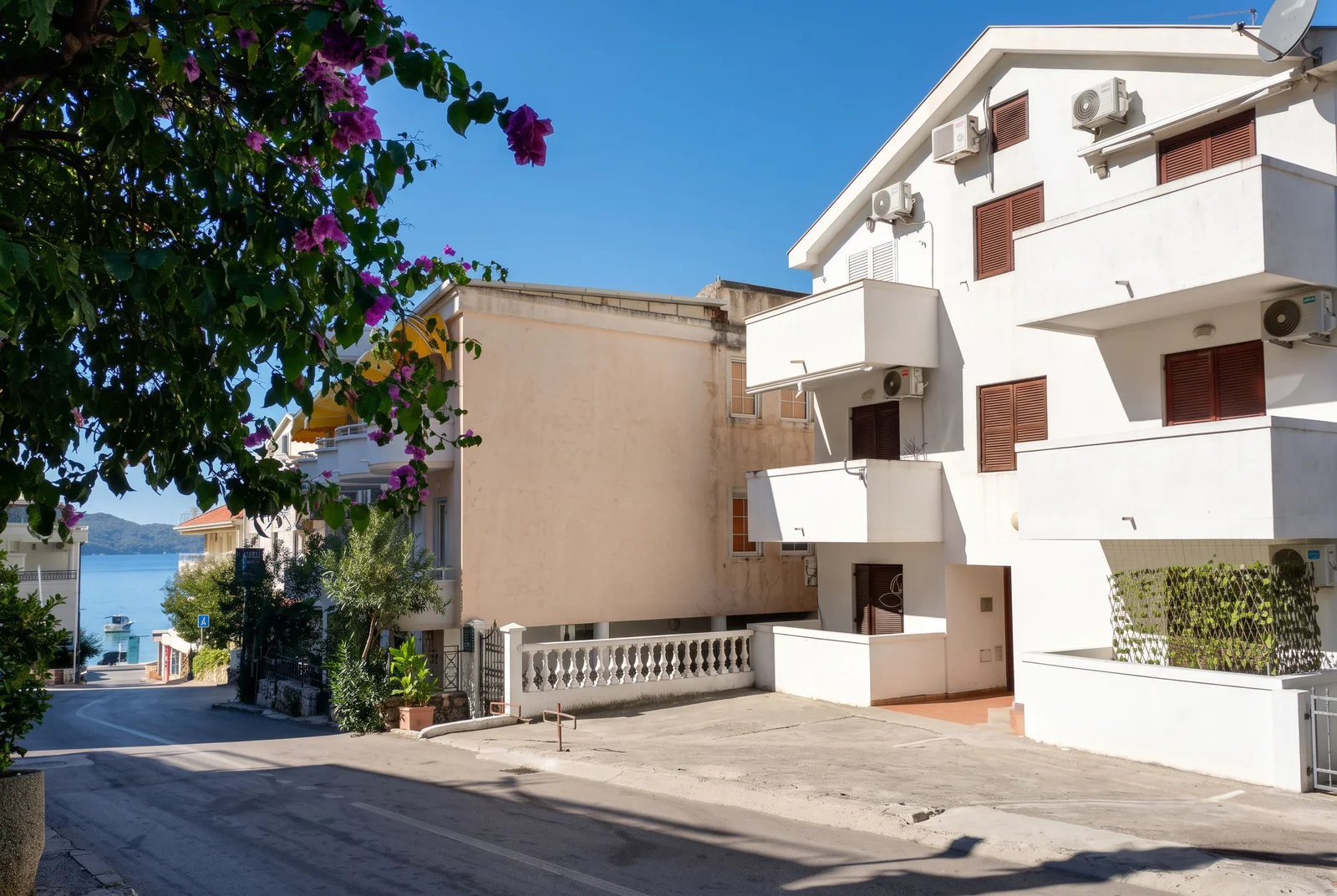 Street view of white residential buildings with balconies and AC units, featuring purple bougainvillea flowers in the foreground.