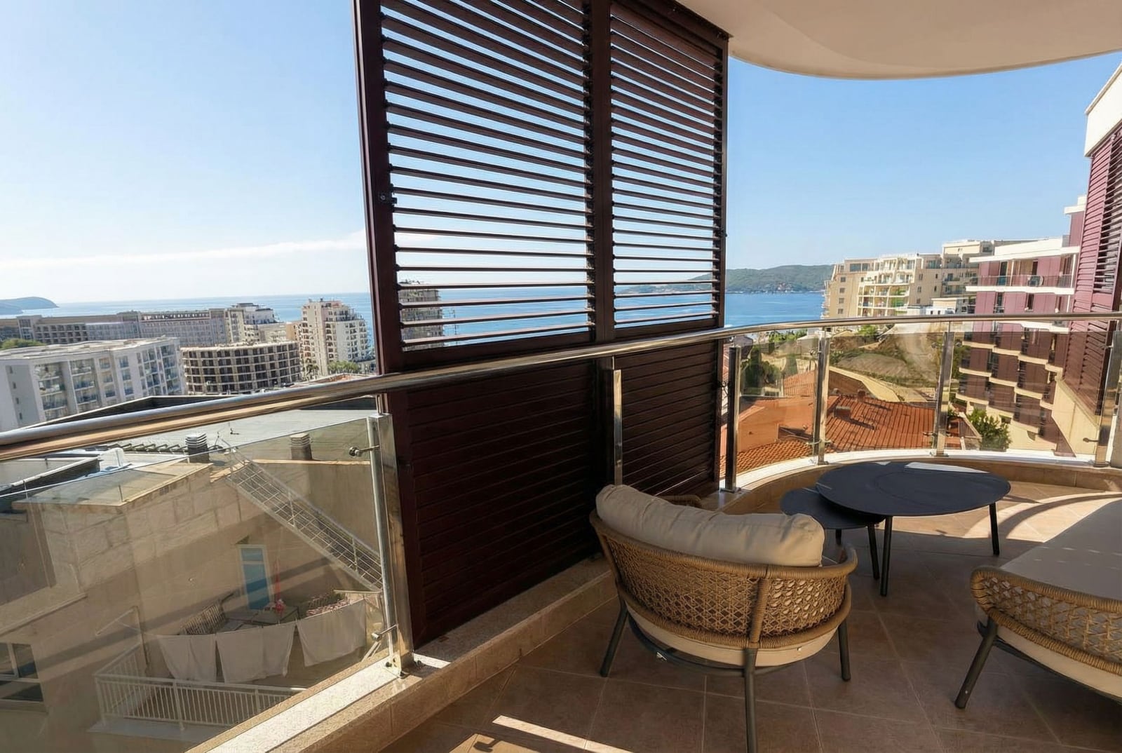 Apartment balcony in Becici, Montenegro: wicker furniture, glass railing, and panoramic views of the Adriatic Sea.