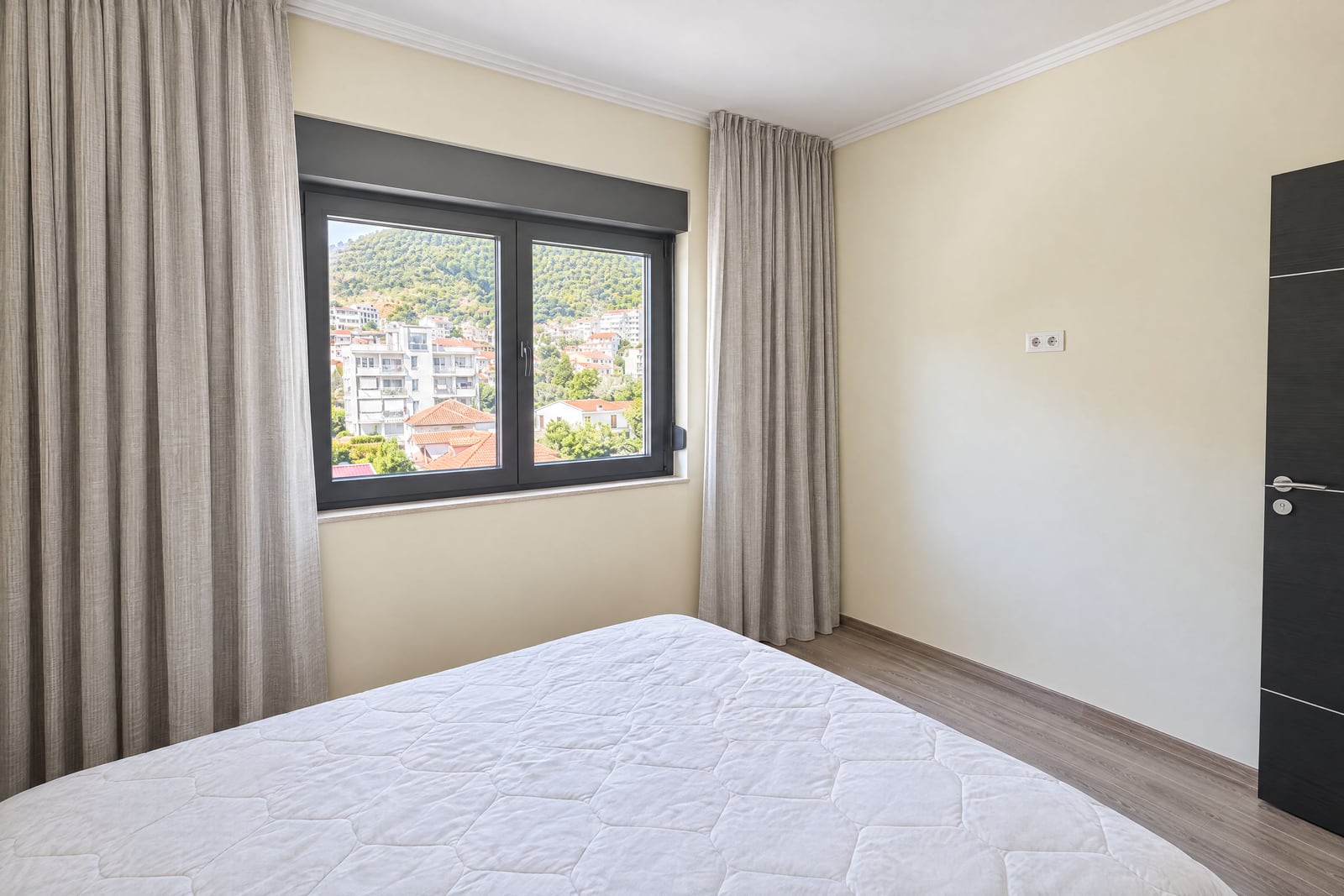 Bright bedroom with a white mattress, beige curtains, and a window overlooking a town and hills.