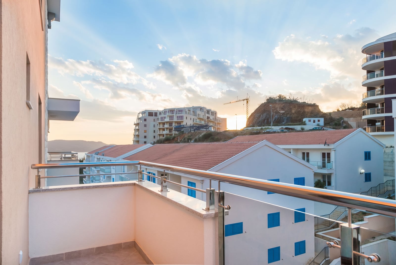 View from the apartment balcony in Bečići, Montenegro: modern houses with tiled roofs, mountains, and a sunset sky.