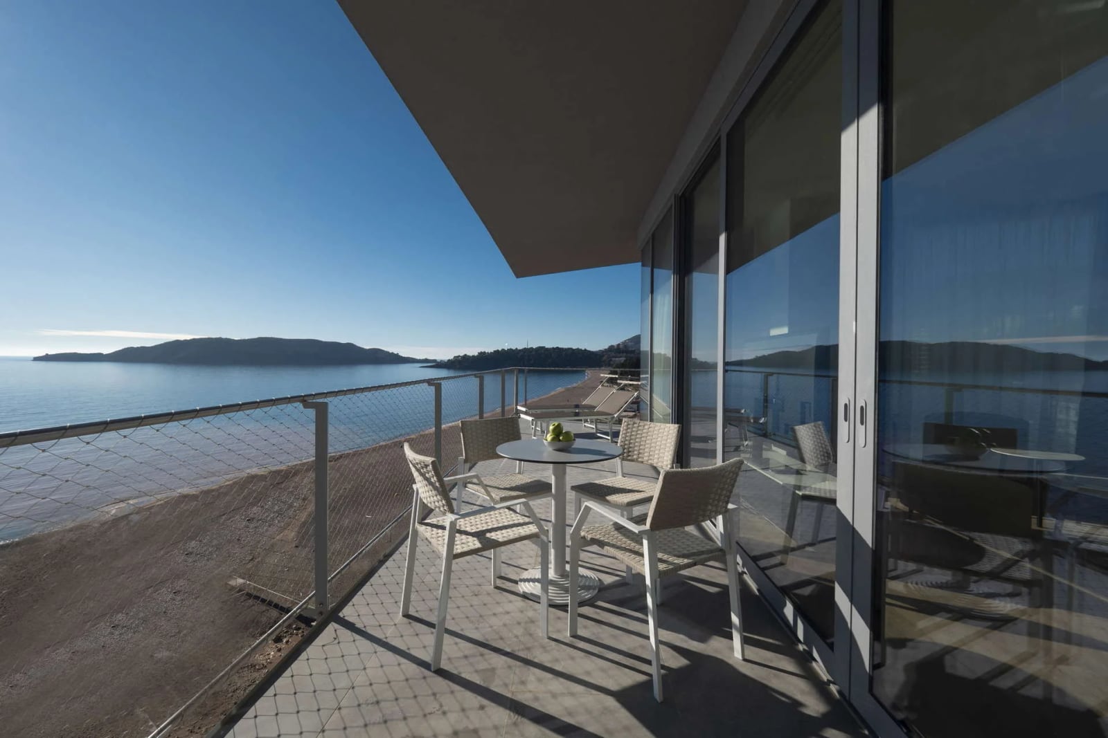 Balcony with white wicker chairs and table overlooking the sea and mountains through glass doors.
