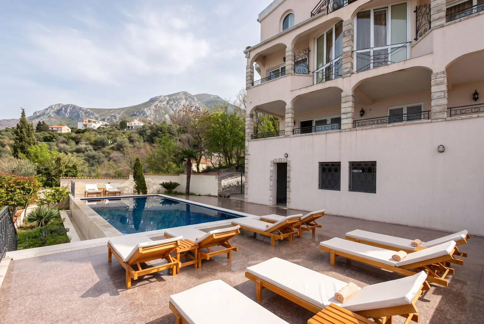 Outdoor swimming pool with wooden sunbeds on a terrace, featuring a mountain view and villa facade.
