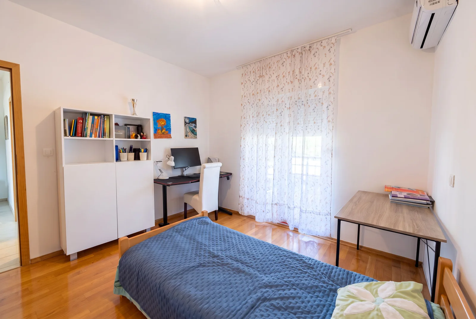 Bedroom featuring a bed with blue cover, white bookshelf, computer desk, and wooden flooring.