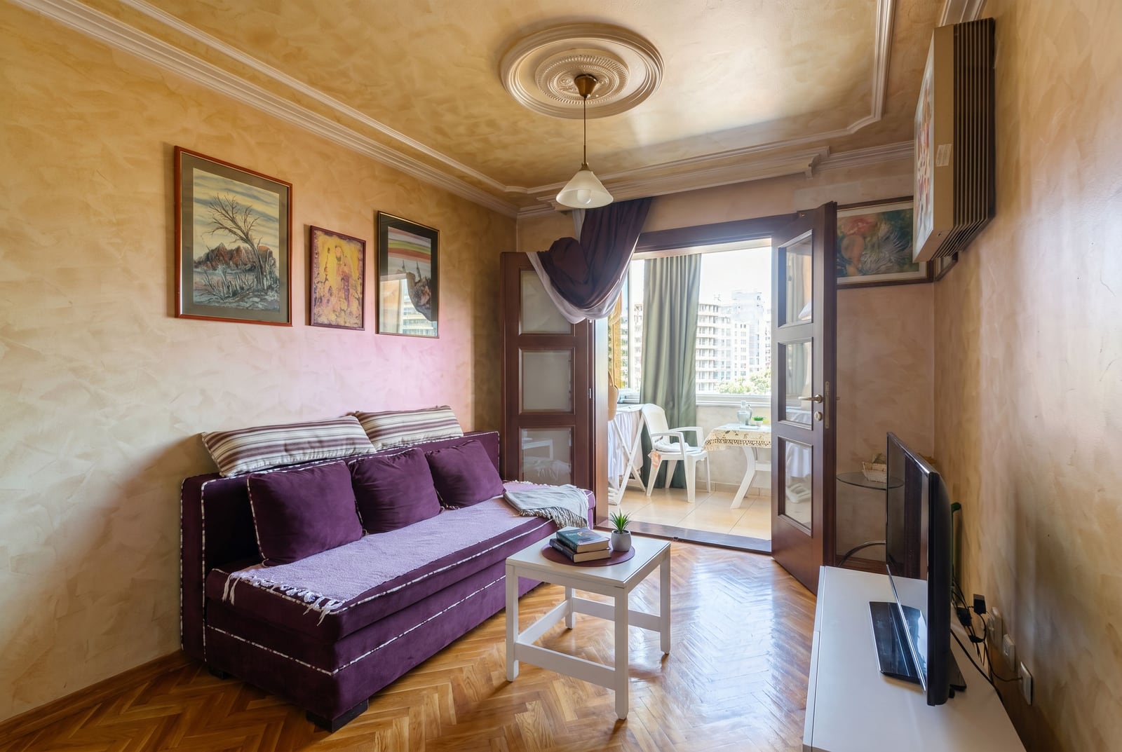 Cozy living room in a Budva apartment, Montenegro: purple sofa, parquet flooring, and balcony access with a city view.