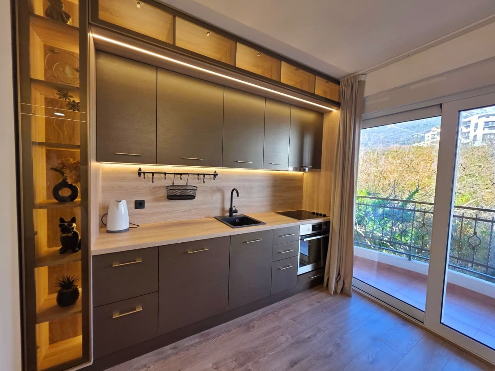 Modern kitchen in an apartment in Bečići, Montenegro: dark cabinets, built-in appliances, and access to a balcony with a view.