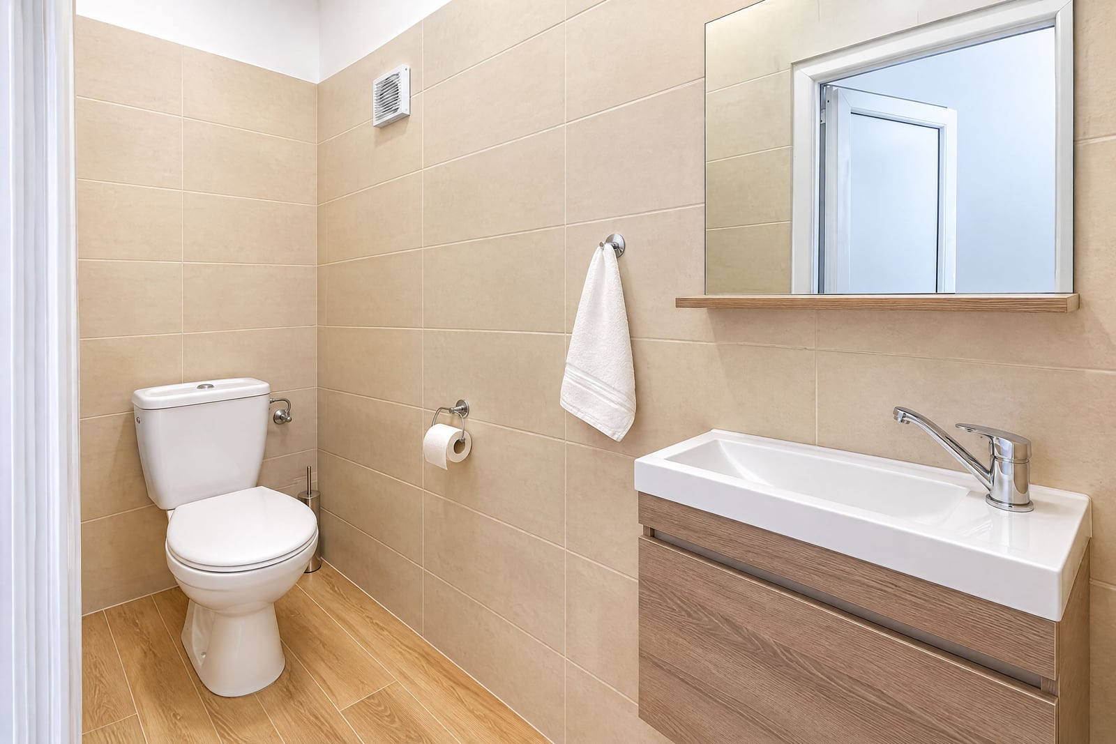 Modern bathroom featuring a white toilet, wooden vanity sink, wall mirror, beige tiled walls, and wood-look flooring.