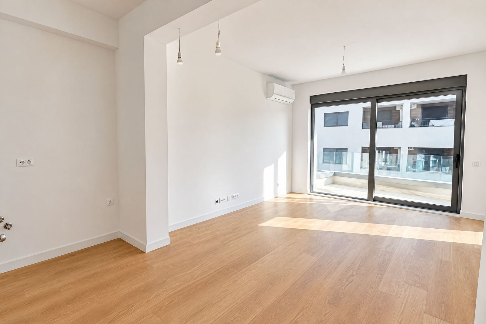 Empty room with wooden flooring, white walls, pendant lights, and a large sliding glass door leading to a balcony.