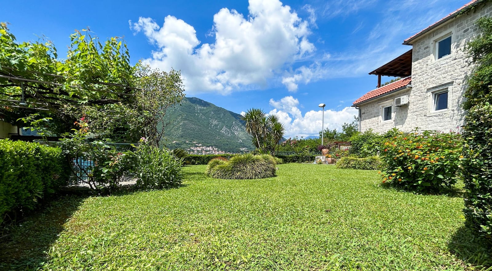 Green garden with a stone villa in Dobrota, Montenegro, with mountain views and a grape pergola on the property.
