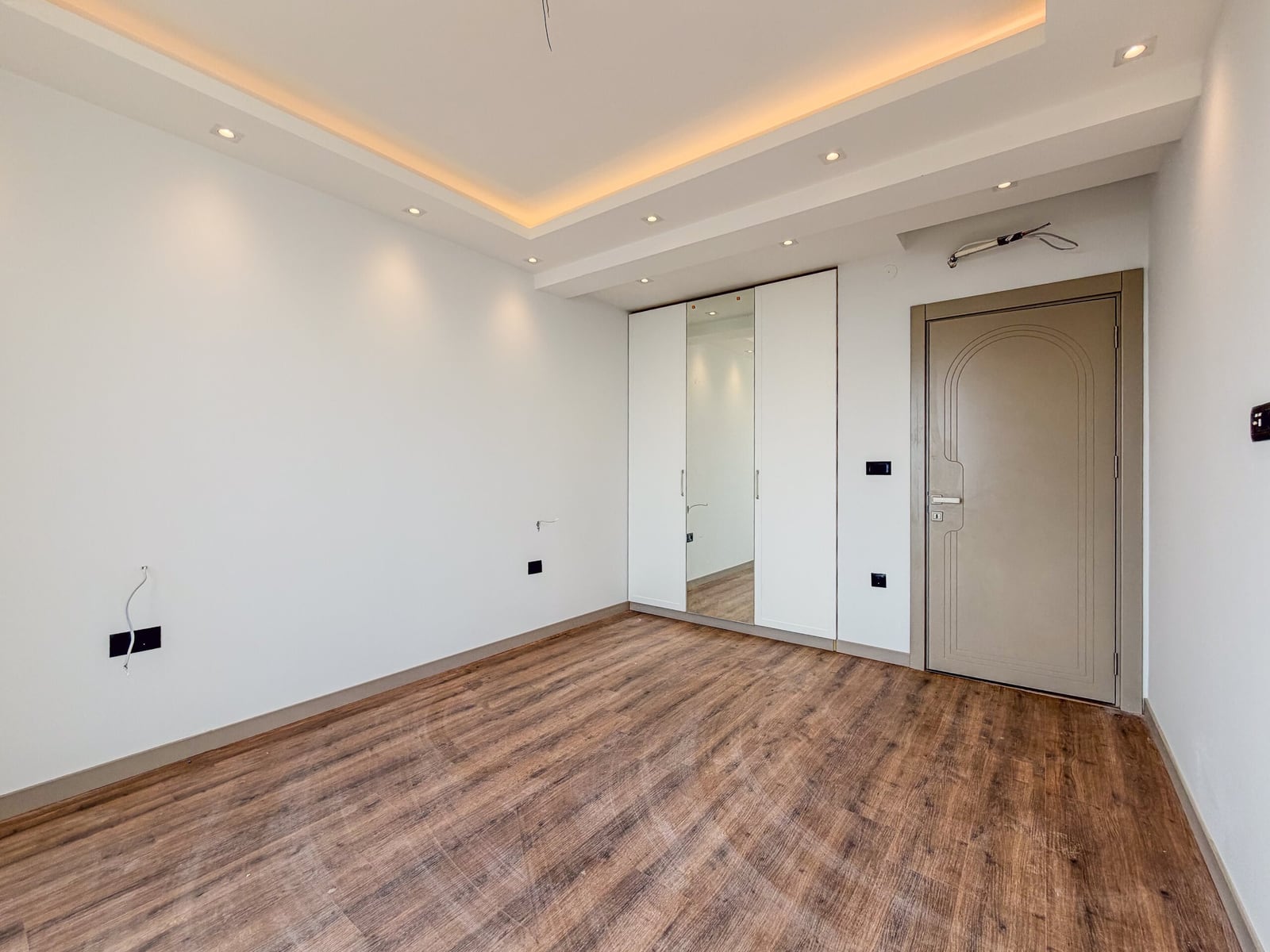 Empty room with laminate flooring, built-in wardrobe with mirror, entrance door, and exposed wiring.
