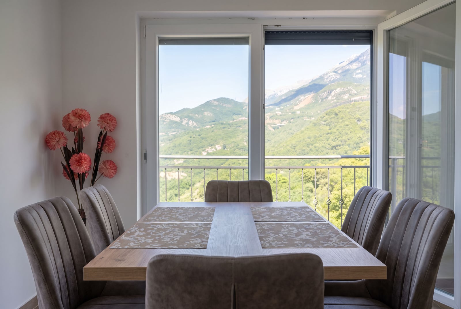 Dining area with a wooden table and upholstered chairs against a panoramic window with mountain views in an apartment, Bečići, Montenegro.