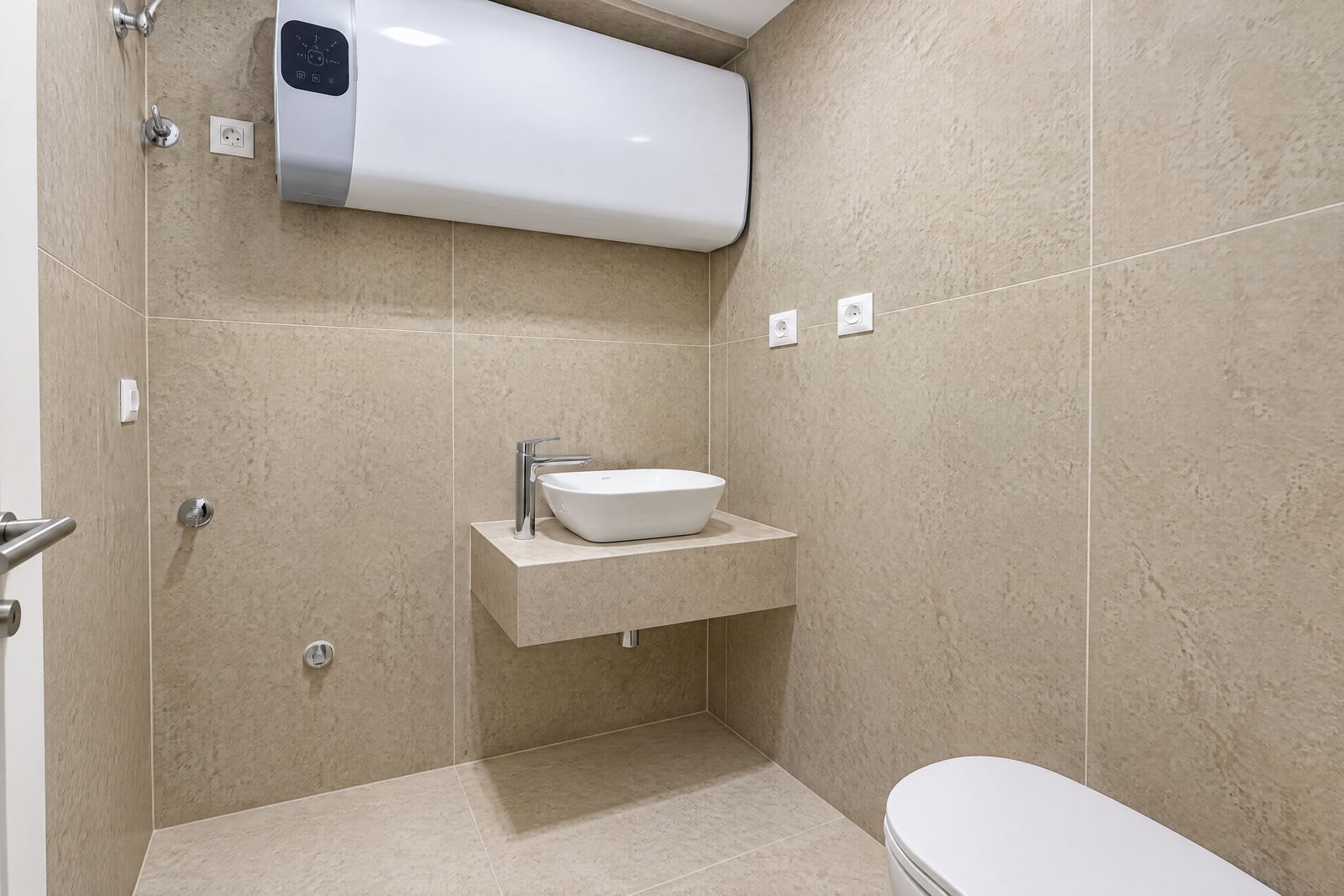 Modern bathroom with beige tiles, a vessel sink on a floating shelf, toilet, and wall-mounted water heater.
