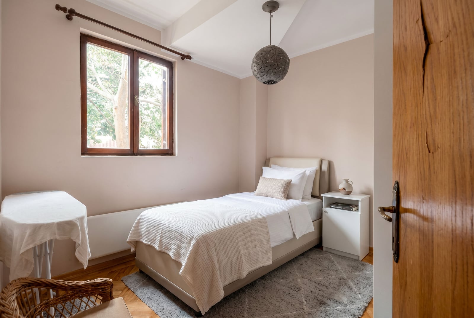 Cozy bedroom with a single bed in a Budva apartment, Montenegro: bright interior, parquet flooring, and a view from the window.