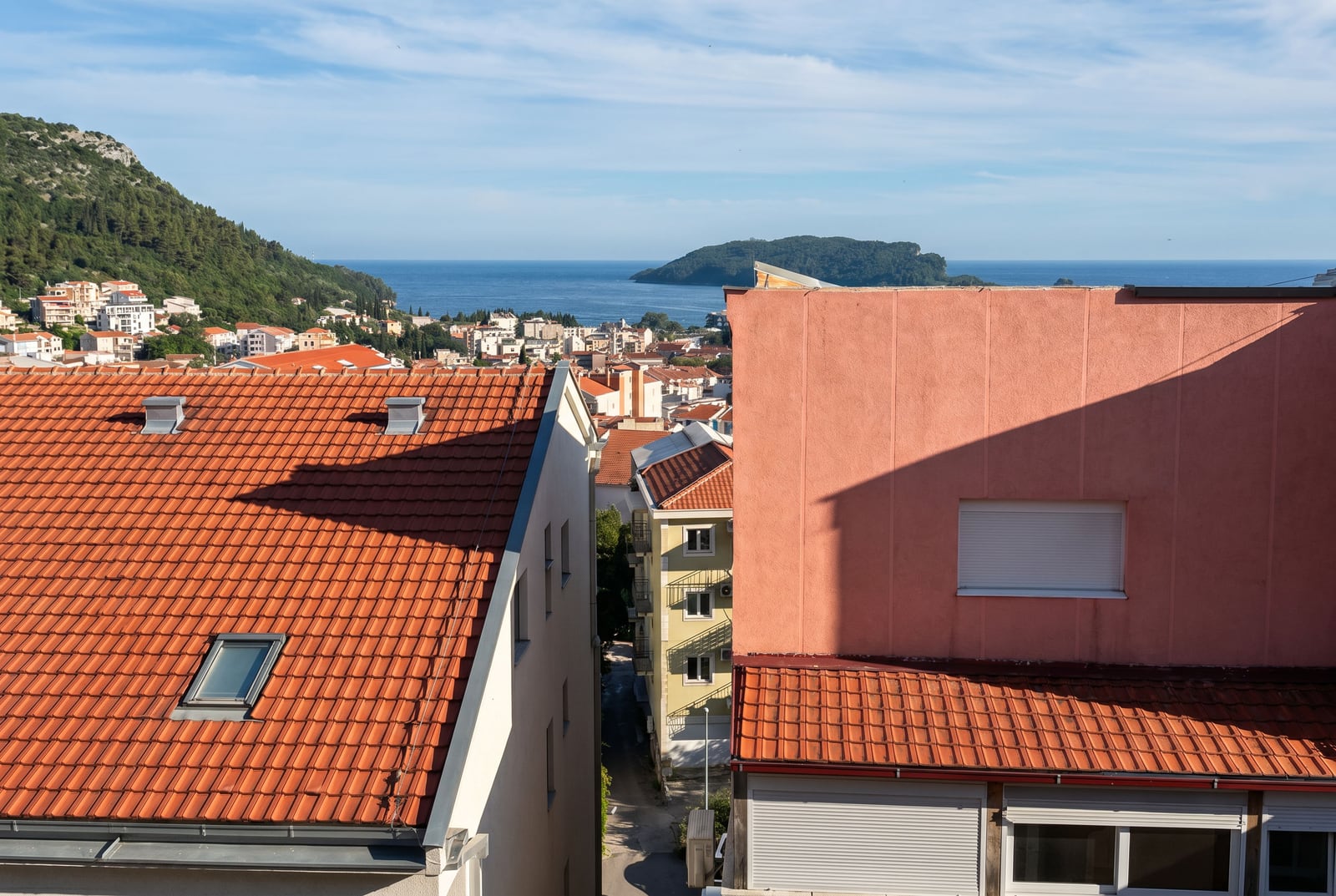 View of the sea and the island of Sveti Nikola from an apartment window in Budva, Montenegro, with red-tiled rooftops in the foreground.