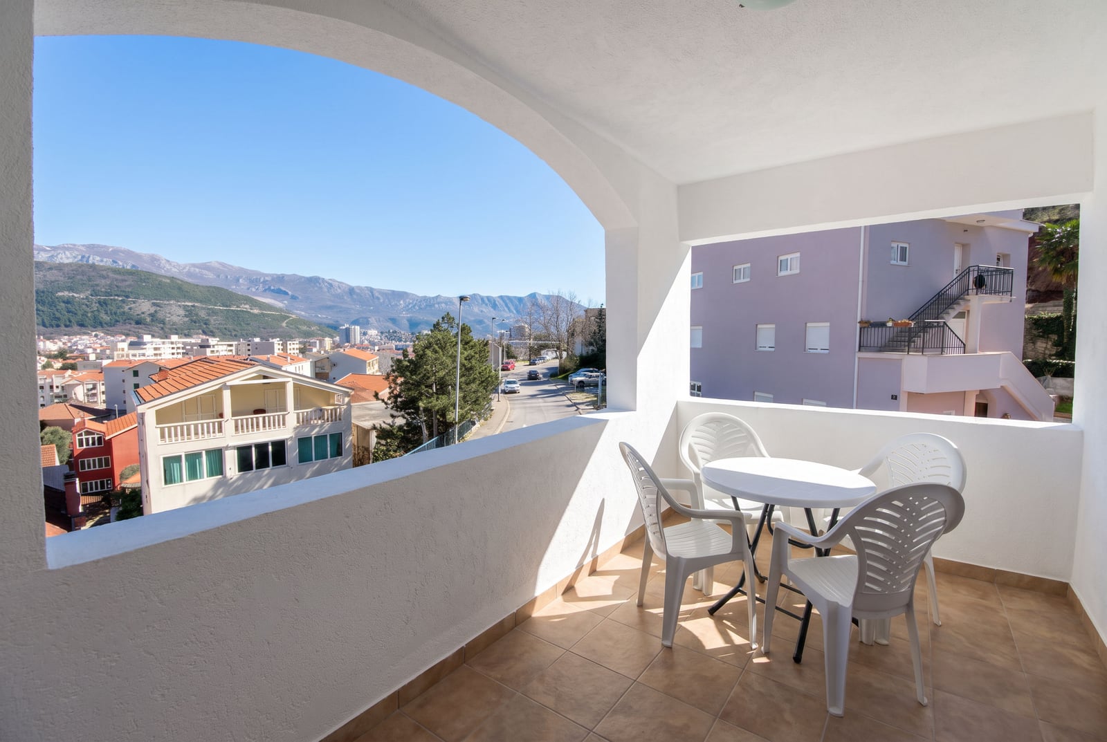 Balcony with white plastic furniture and arched opening overlooking a town and mountains.