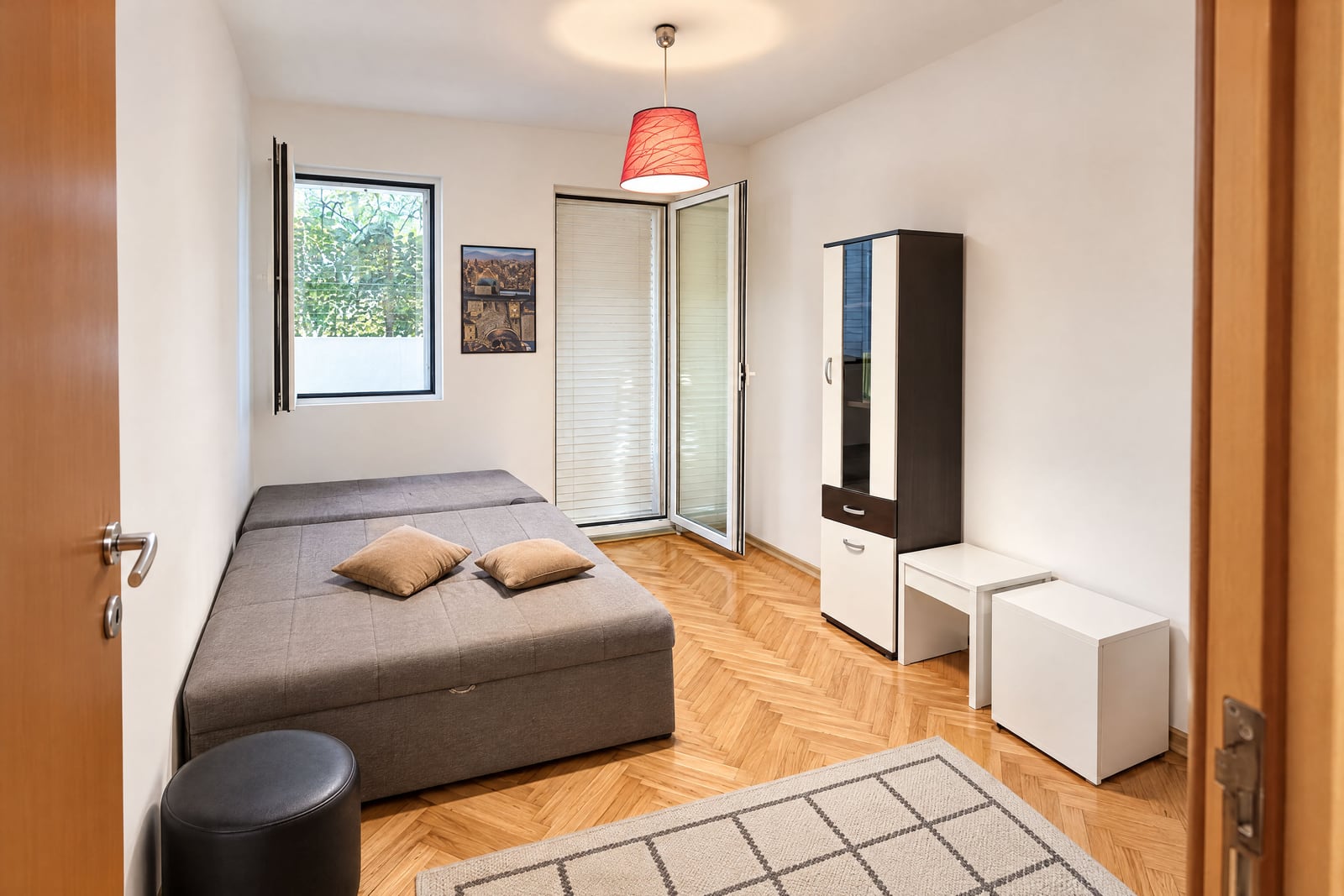 Bedroom featuring a grey sofa bed, parquet flooring, a tall storage cabinet, and a red pendant lamp.