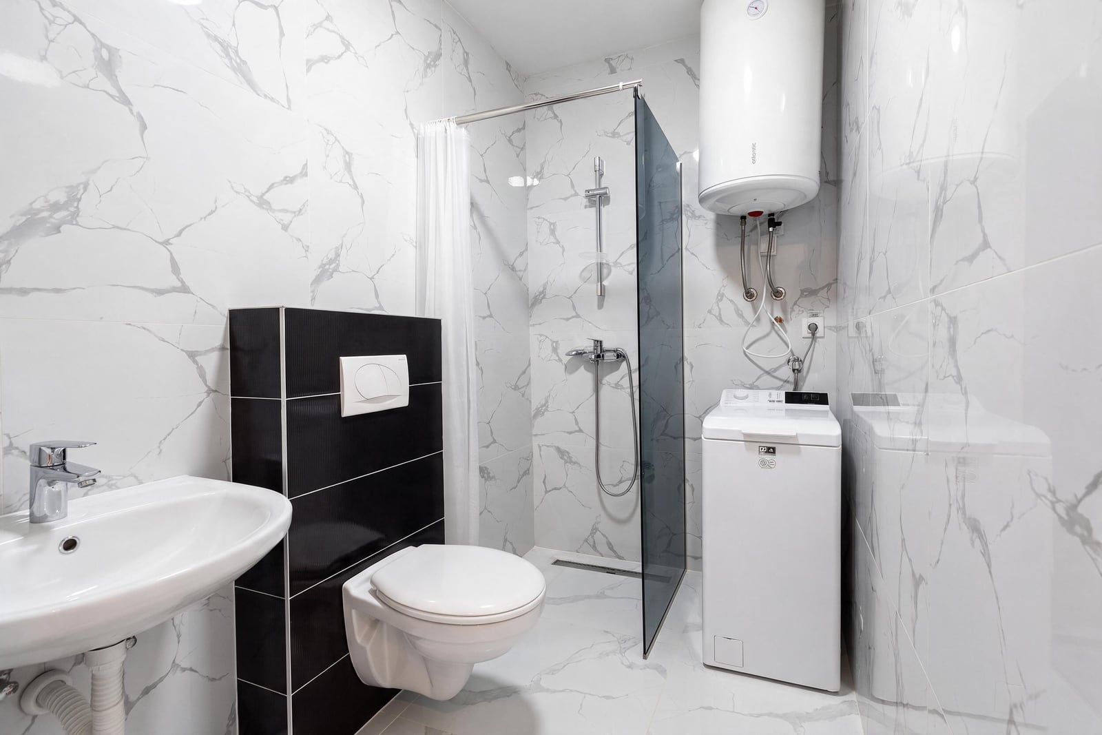 Bathroom featuring a white toilet, sink, shower area with glass partition, washing machine, and marble-effect tiles.