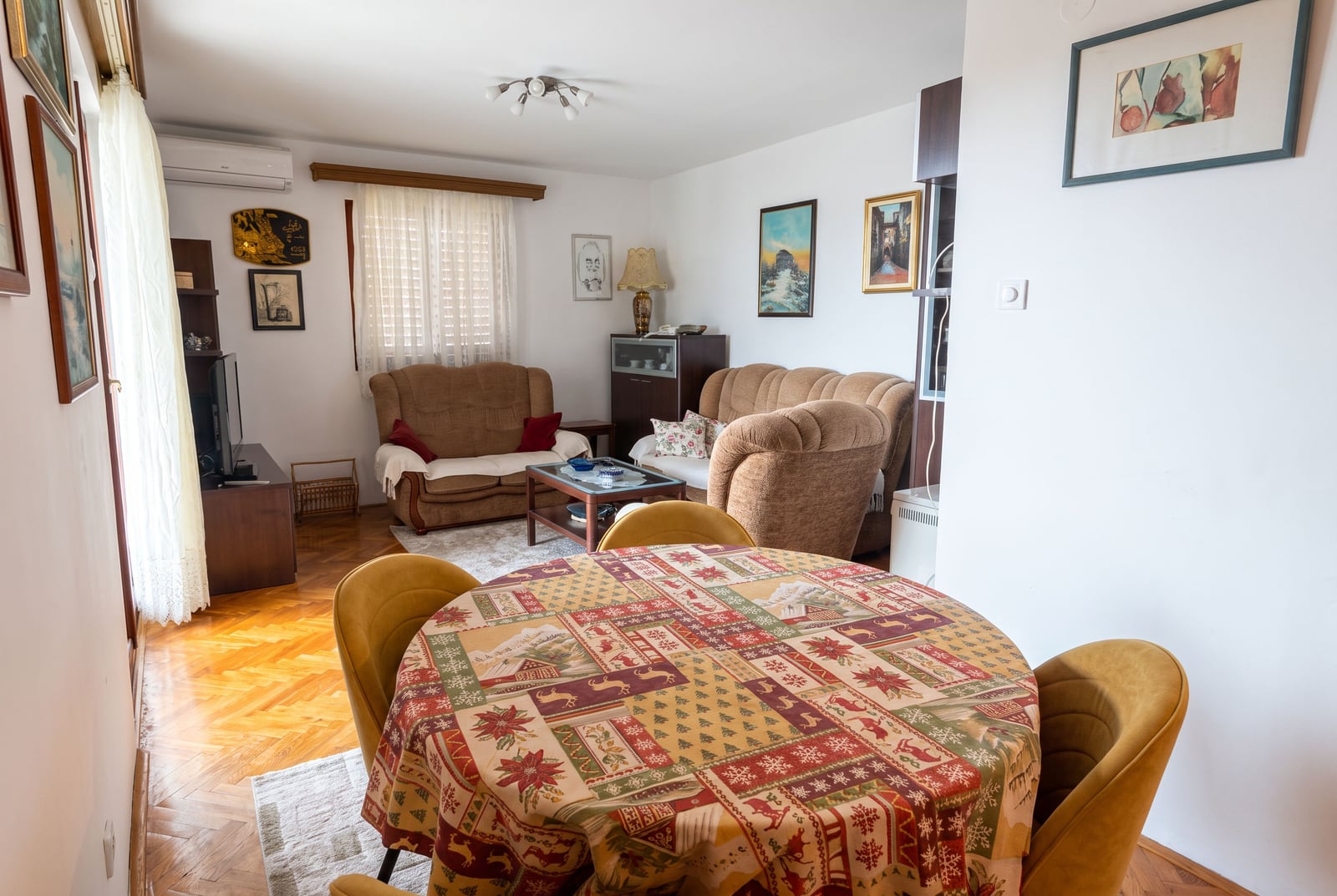 Living room featuring brown sofas, a coffee table, and a round dining table with a patterned cloth on parquet flooring.