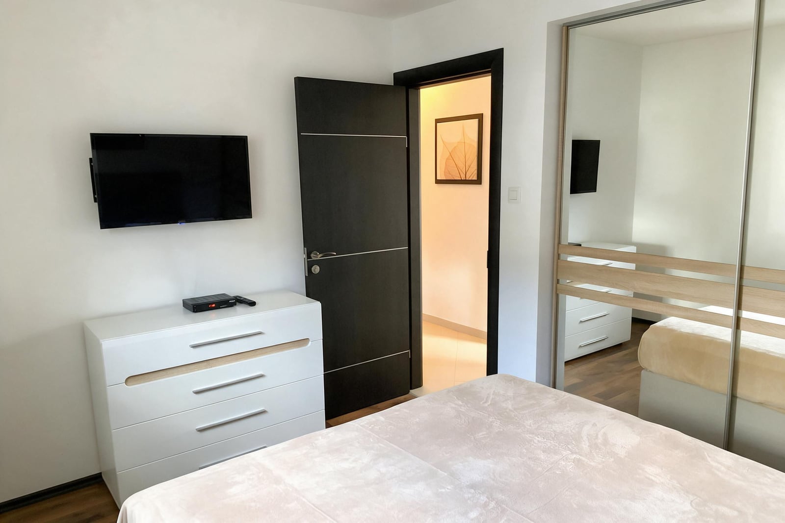 Modern bedroom featuring a white dresser, wall-mounted TV, and large mirrored wardrobe closet.