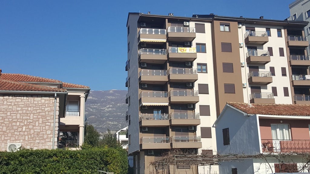 Modern multi-apartment building with balconies in Budva, Montenegro, against a backdrop of mountains and clear sky.