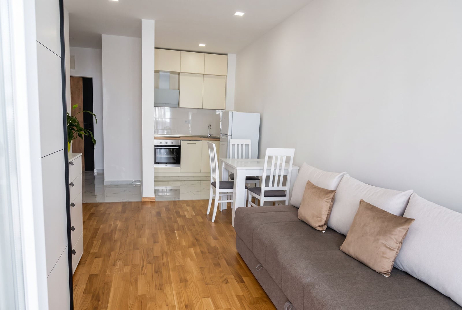 Living room with a grey sofa and wooden floor; an open kitchen with white cabinets and a dining table is visible in the background.