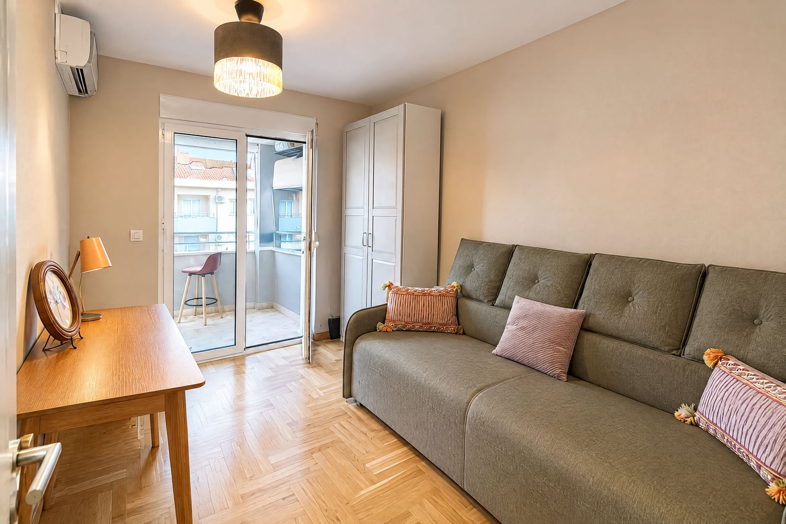 Living room with a grey sofa, wooden table, white wardrobe, and parquet flooring leading to a balcony.