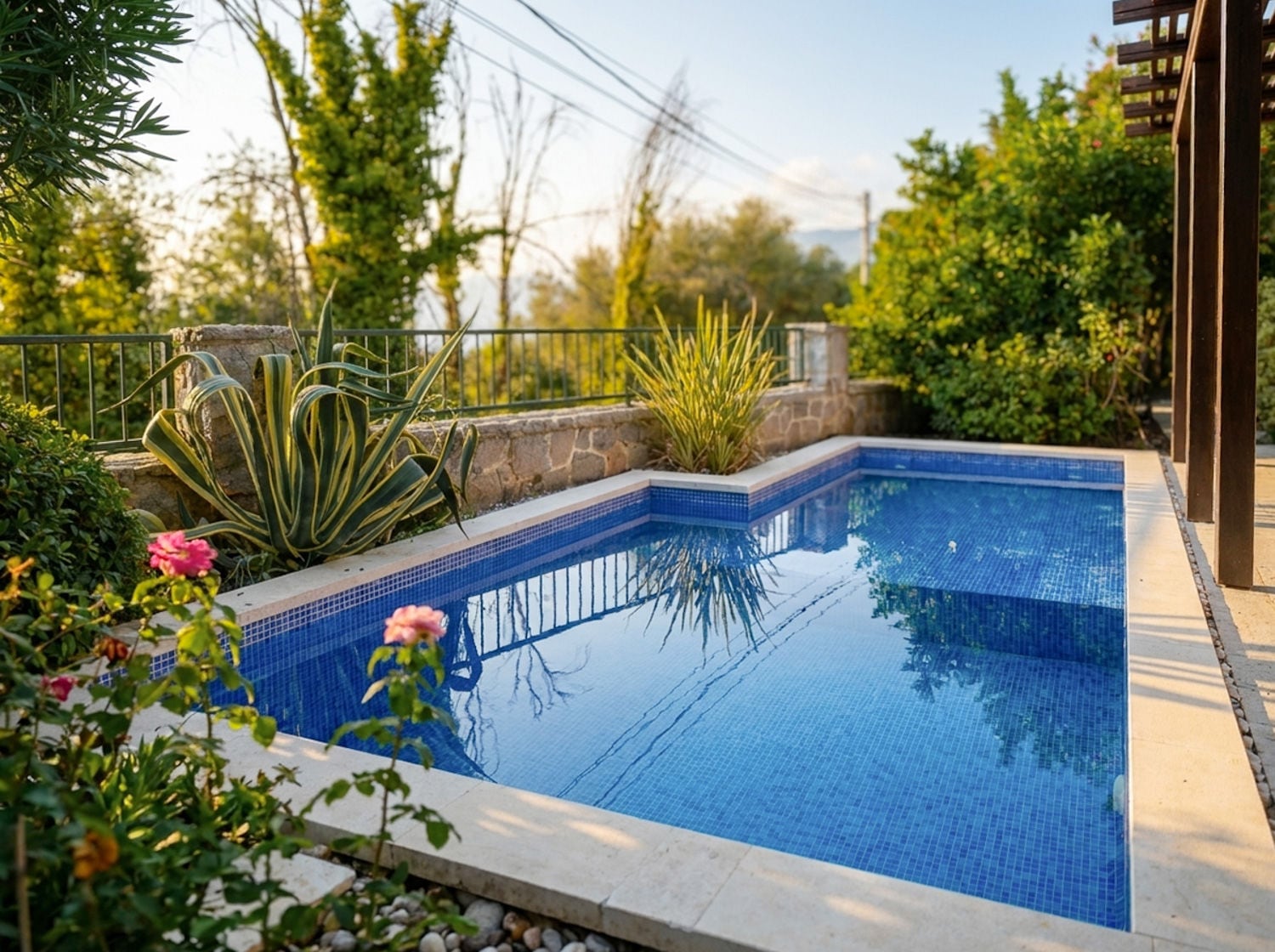 Small blue tiled outdoor swimming pool surrounded by lush greenery, agave plants, and a stone wall.