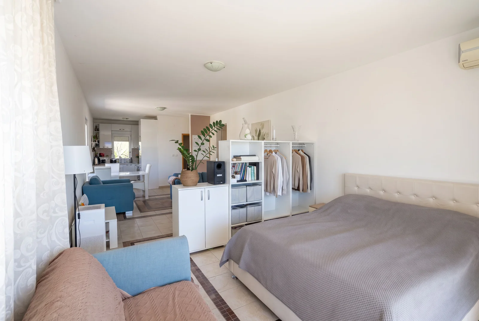 Bedroom featuring a large bed, open clothing rack, blue sofa, and a view into the kitchen area.