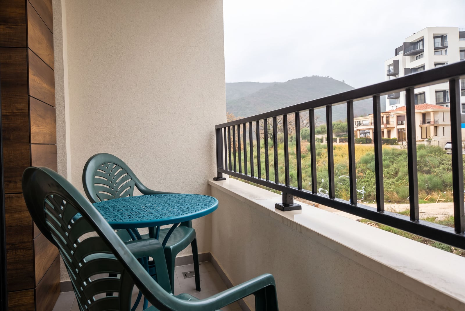 Balcony with green plastic chairs and table, black metal railing, overlooking hills and nearby residential buildings.